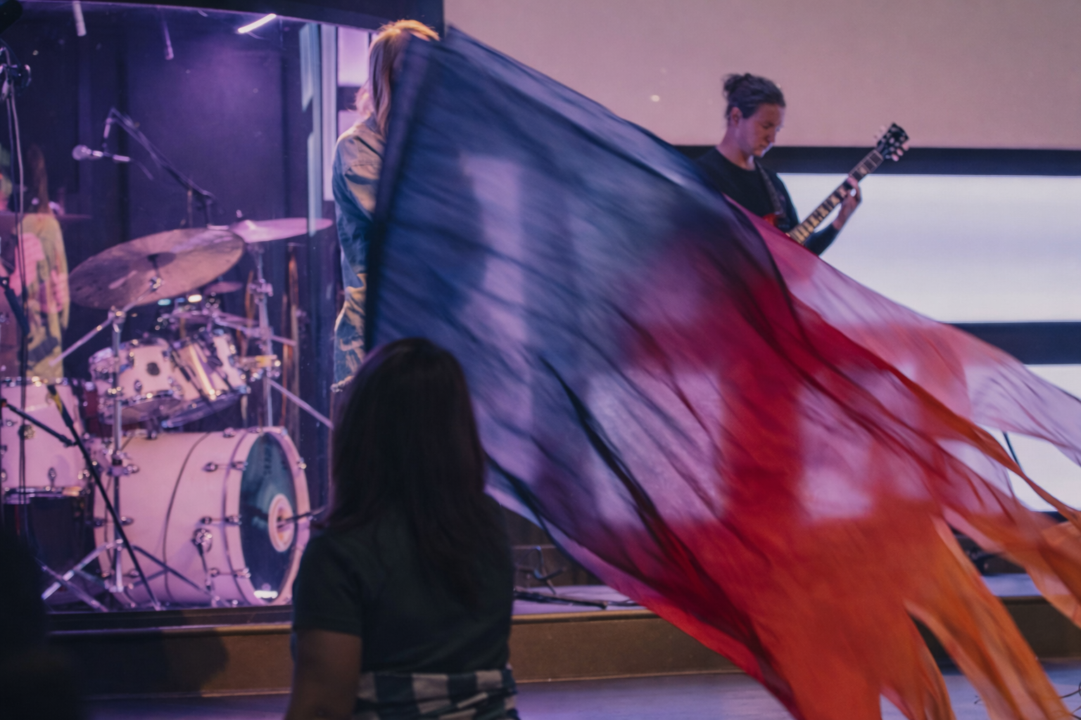 Musicians performing on stage, with one person holding a large flowing flag in red, purple, and blue colors. A drummer and guitarist are visible in the background.