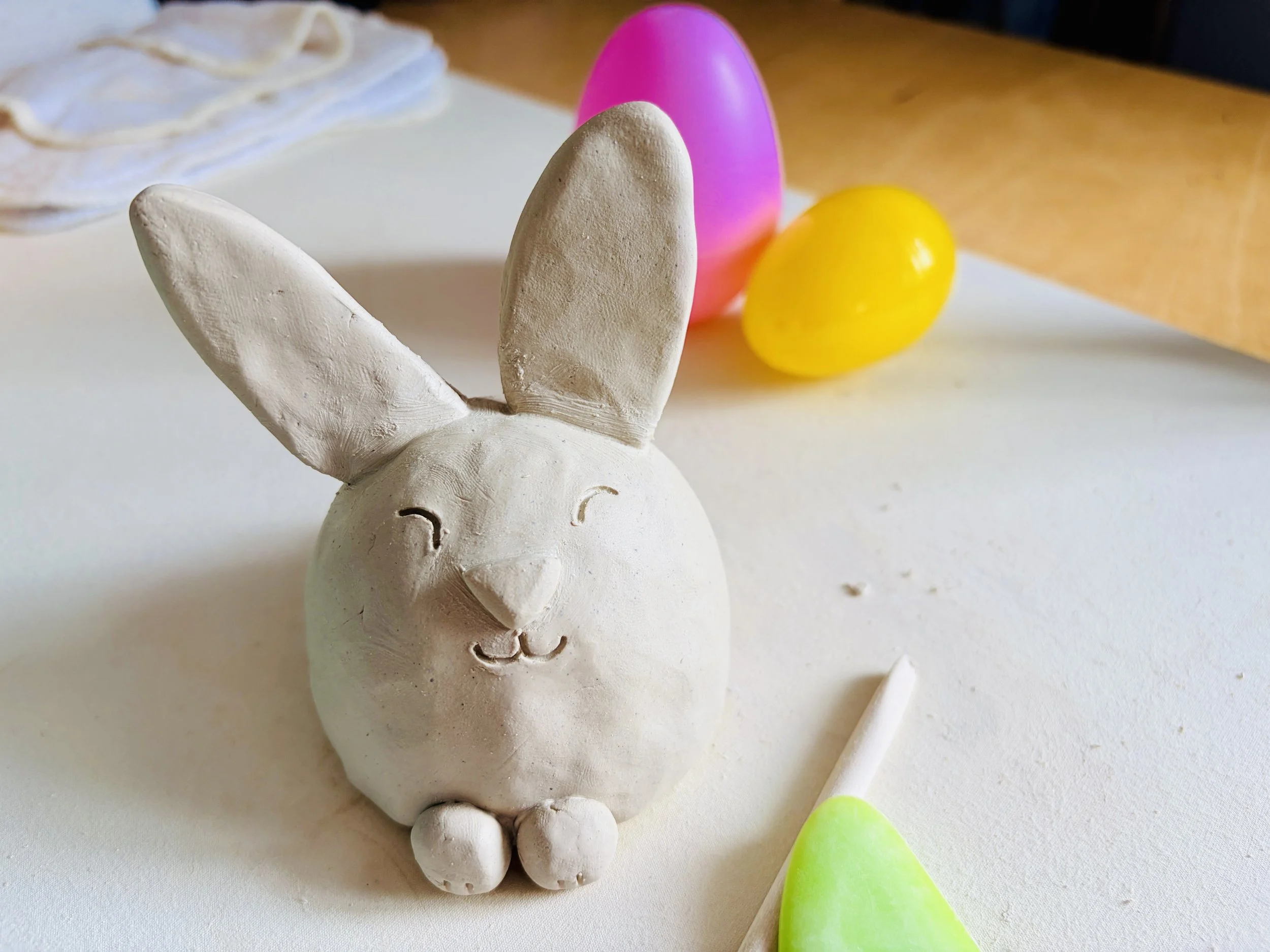 A studio work table with a sculpted clay rabbit that is smiling with two colorful eggs behind it