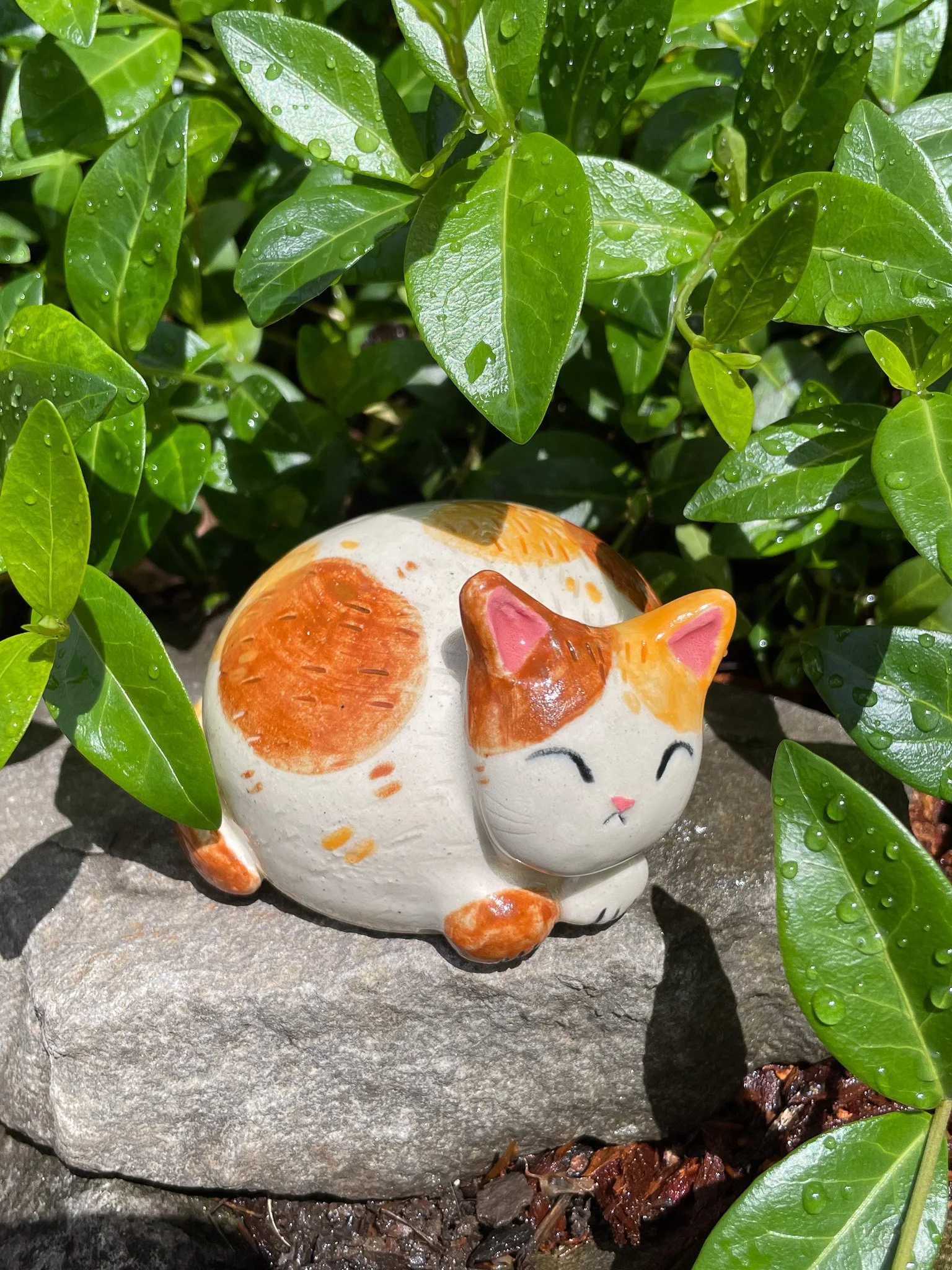 A ceramic figurine of a sleeping cat with orange and white patches, nestled on a gray stone among green leafy plants with water droplets.