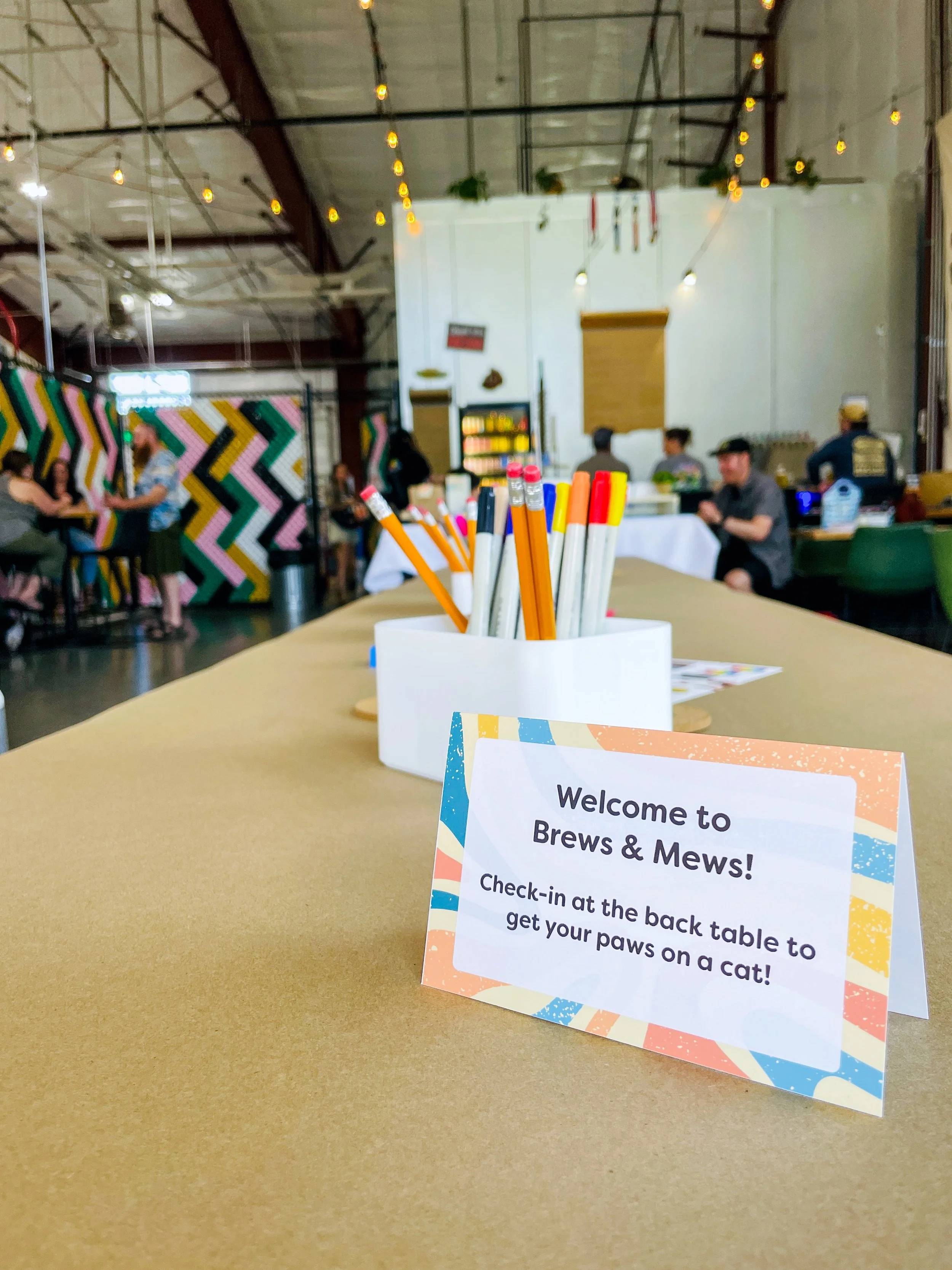 Photo of the inside of the brewery with high ceilings, open space. Customers seated at tables enjoying their beer. Close up of a table with a sign that reads "Welcome to Brews & Mews"