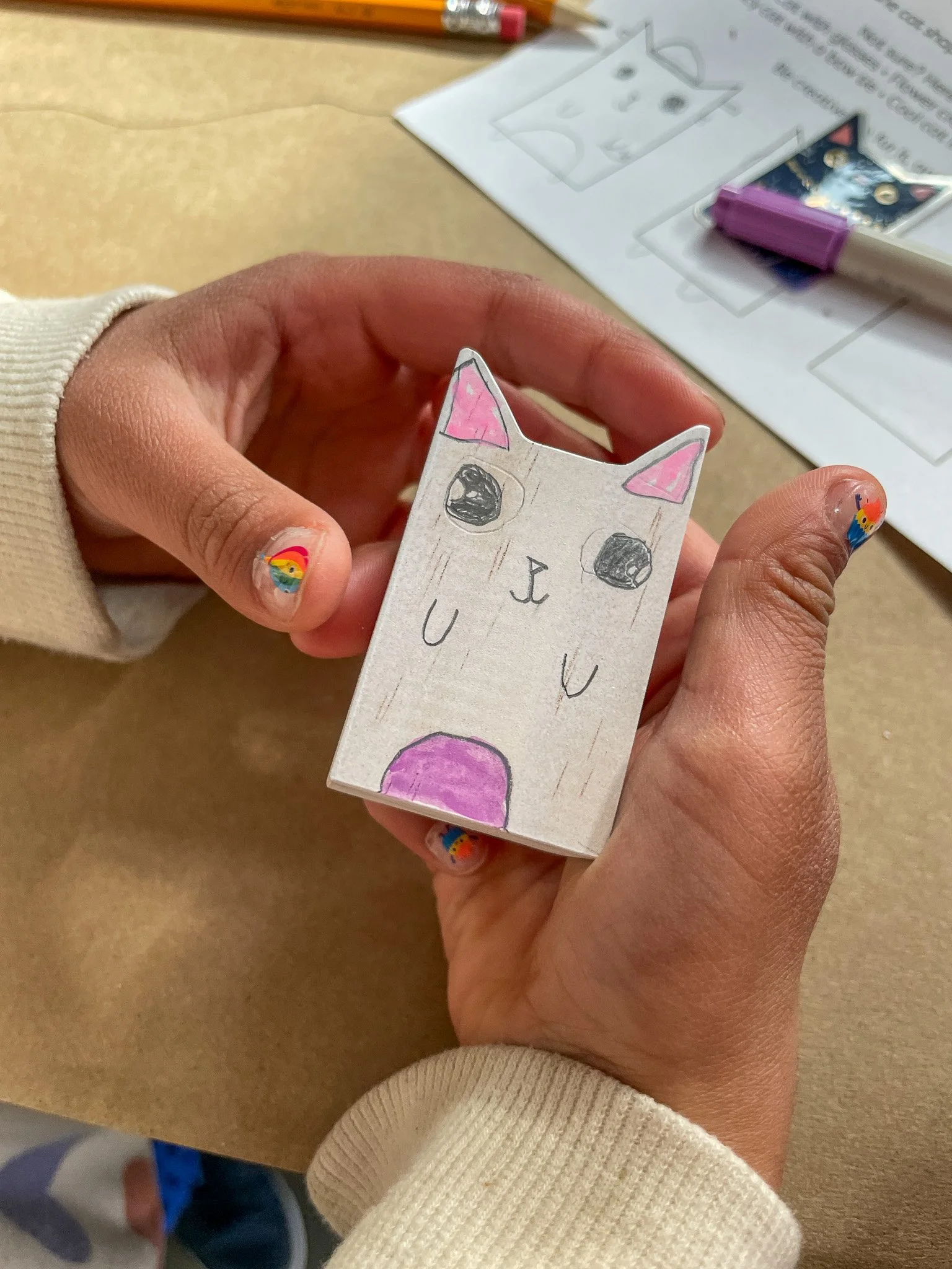 Handmade paper craft depicting a cute, cartoon-like cat with large eyes, pink ears, a small nose, and a purple patch on its lower body, held by a person with rainbow-flower nail art.
