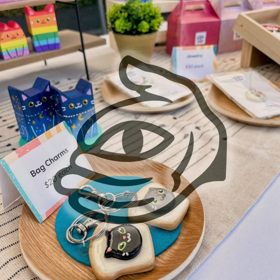 Wooden tray with black cat keychains and stones featuring black cat drawings, beside a sign saying 'Bag Charms $2 each', on a table with decorative items and jewelry display.