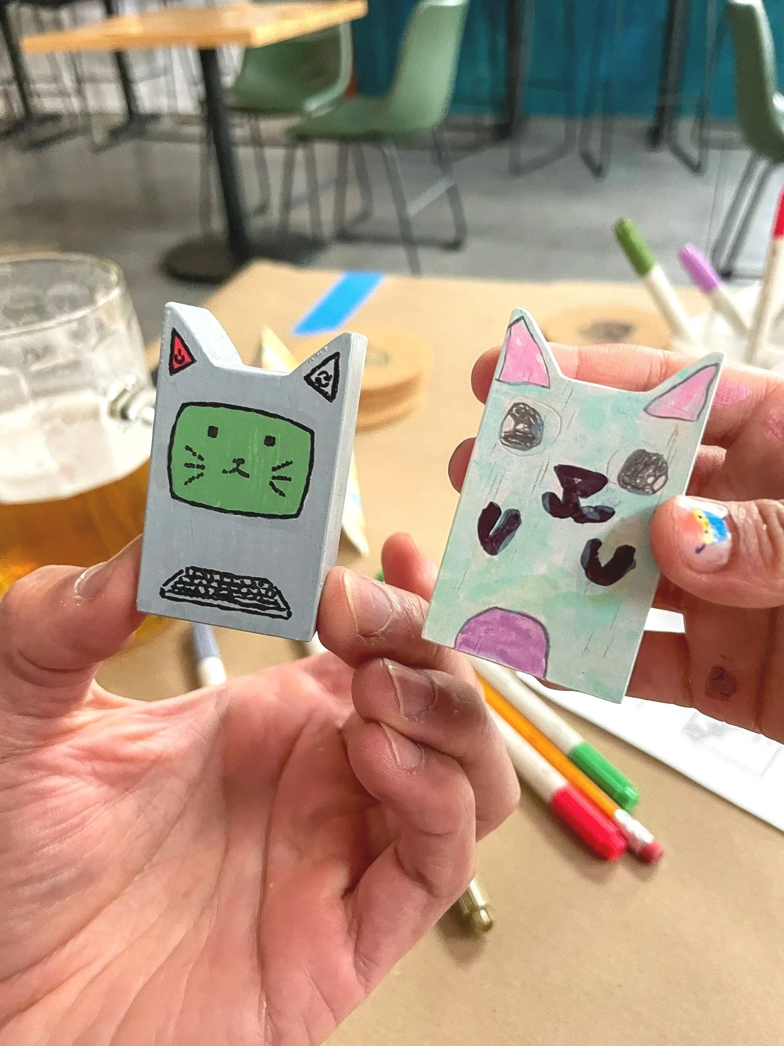 Closeup picture of two people holding their painted miniature cat figurines. One is painted to look like a old computer. The other is a cute pink and purple kitty