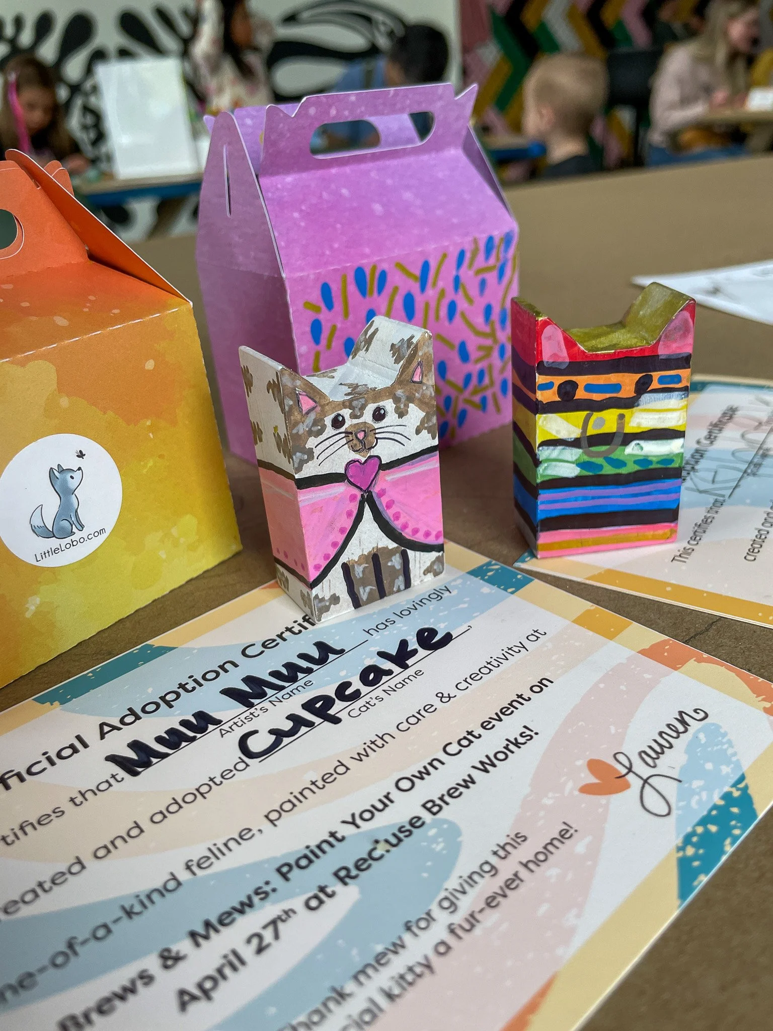 Colorful, hand-painted miniature cardboard boxes and a pink cardboard carrier on a table, with children in the background.