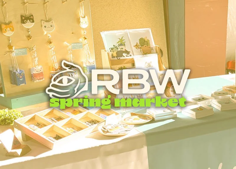 A display table at a spring market with framed artwork, jewelry, and small craft items, with a logo reading "RBW spring market" overlayed.