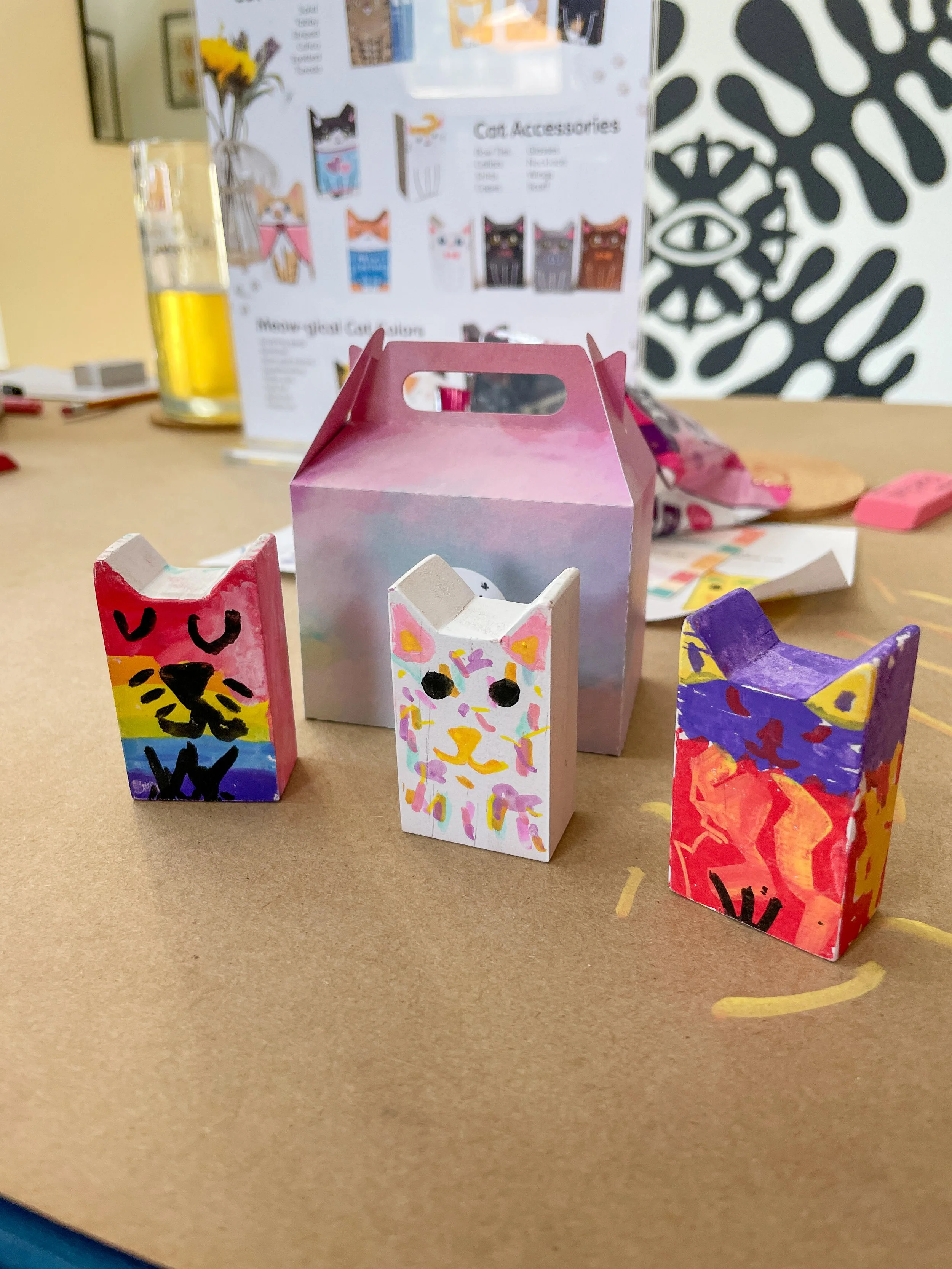 Three painted cardboard cat-shaped containers with different colorful designs, placed on a table with a pink and purple pastel gift box behind them, and an informational display in the background.