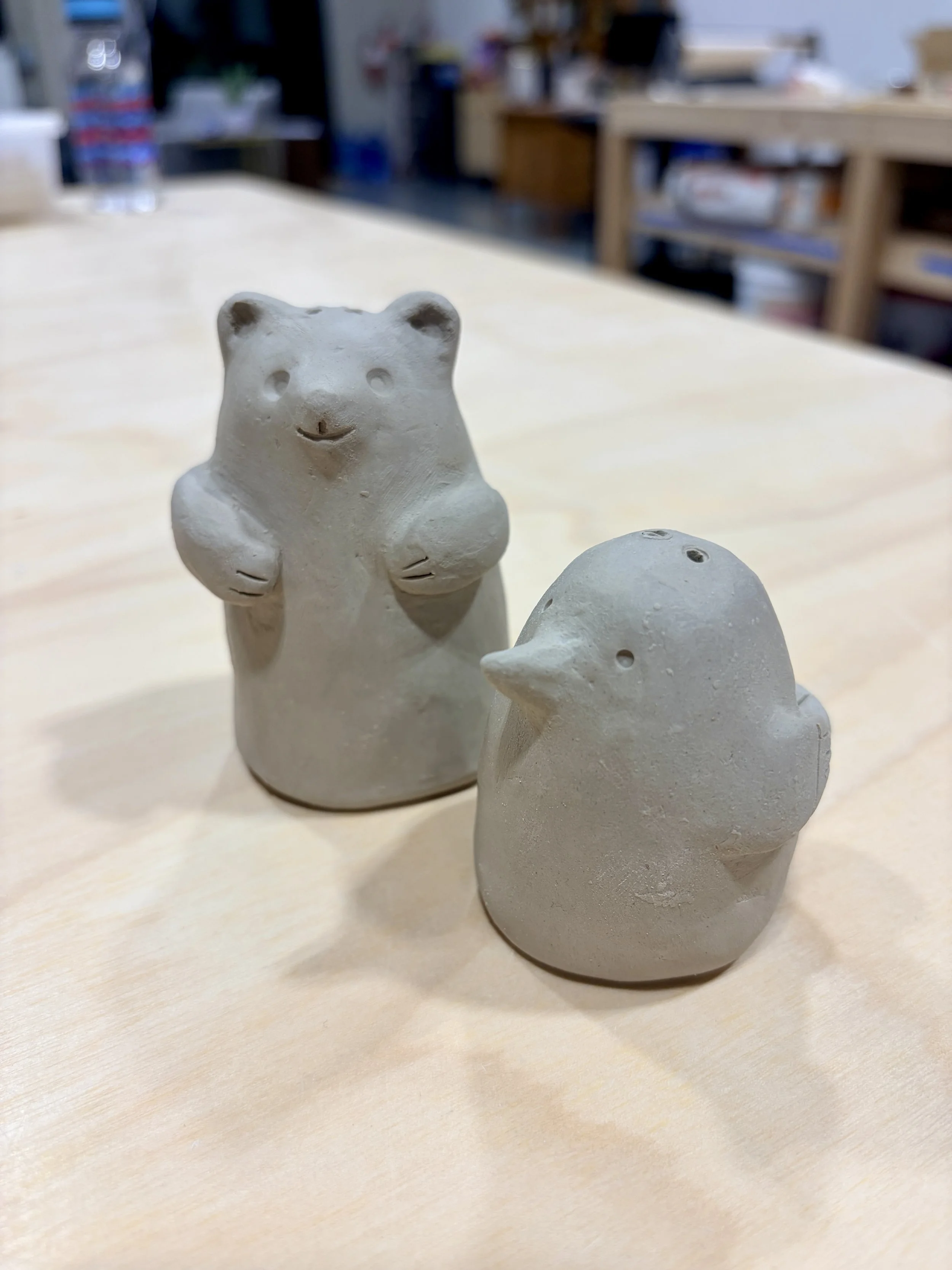 Two small ceramic animal figurines, one shaped like a bear and the other like a duck, on a wooden table in a workshop or studio setting.