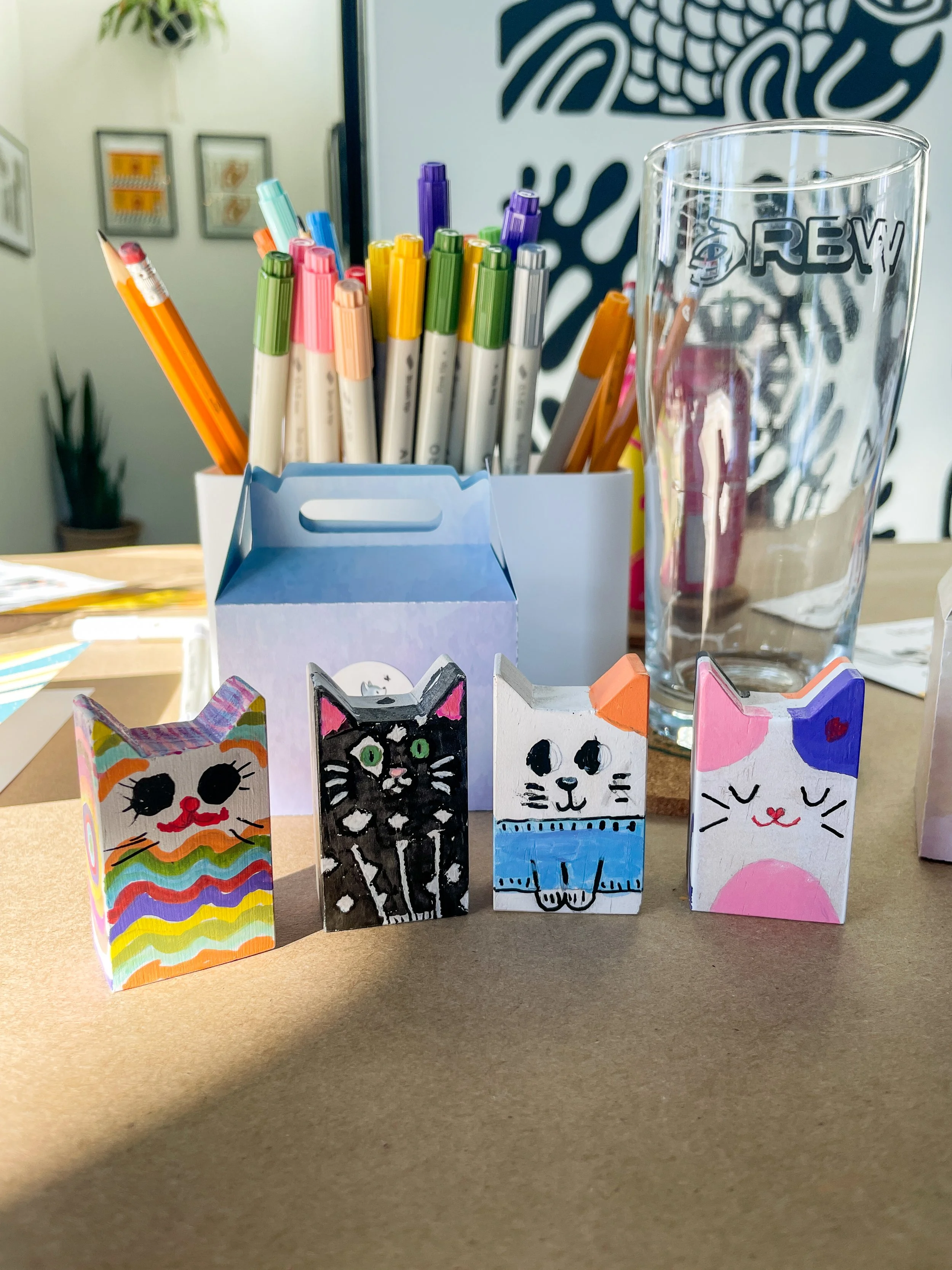 Colorful hand-painted paper cat figurines lined up on a table in front of a holder filled with colored pens and a tall glass.