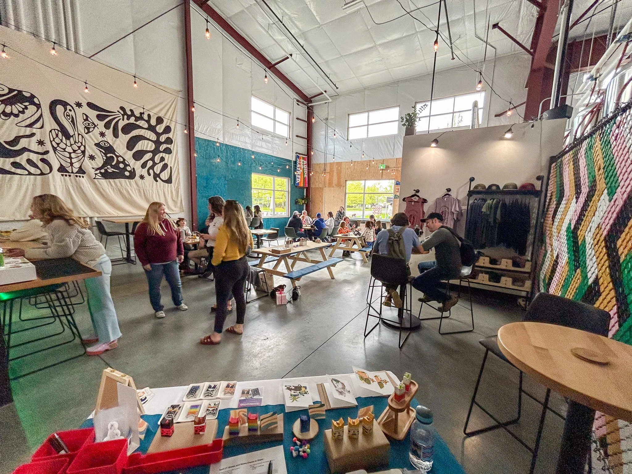 People gathered inside a brightly lit, spacious industrial-style event space with high ceilings, large windows, and string lights. Some are conversing at tables, while others browse merchandise and displays on tables and shelves.