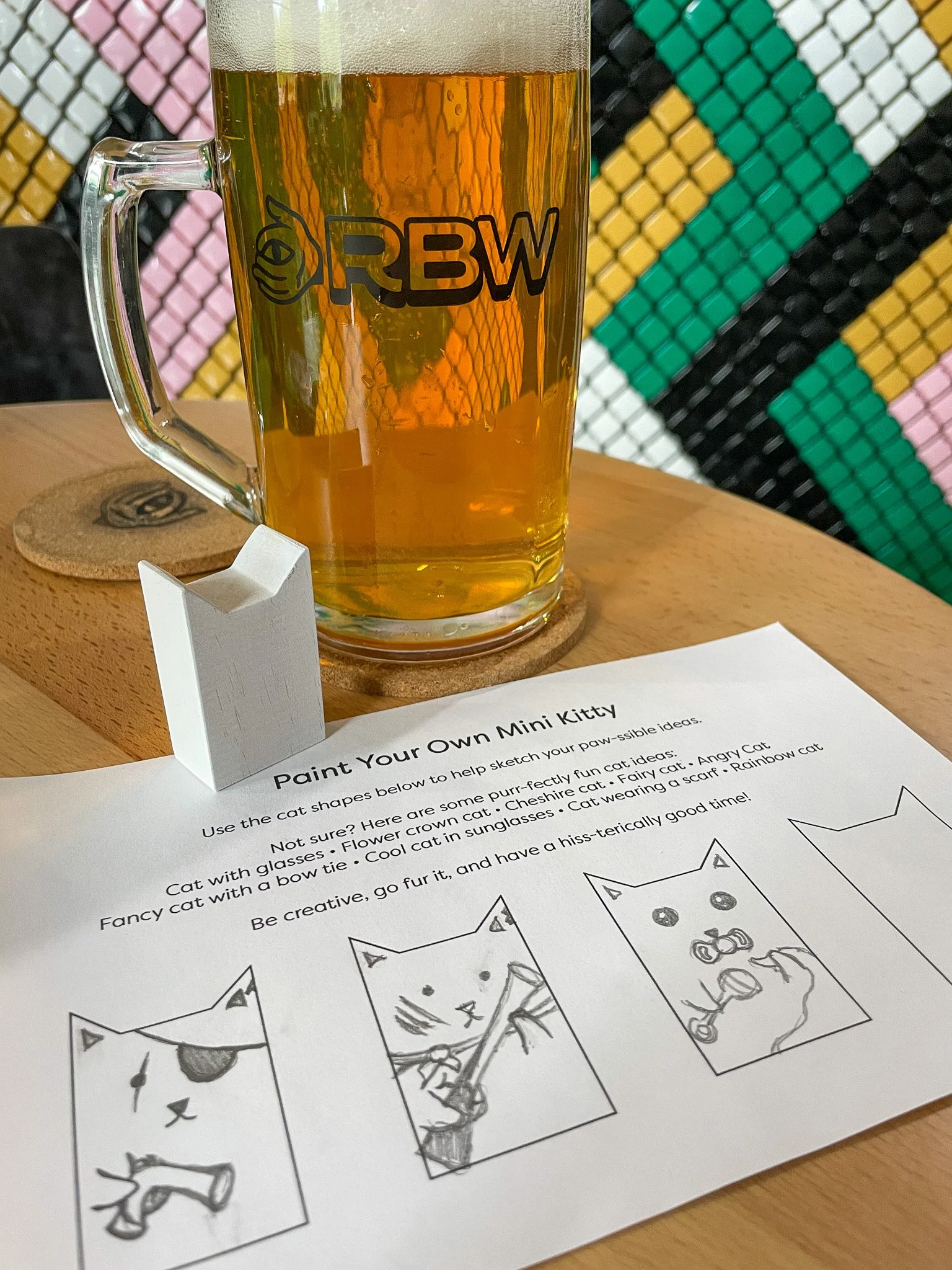 A glass of beer with a logo reading RBW on it, placed on a coaster at a table, with a paint your own mini kitty craft activity sheet underneath it. The activity sheet includes three illustrated steps for drawing a cat.