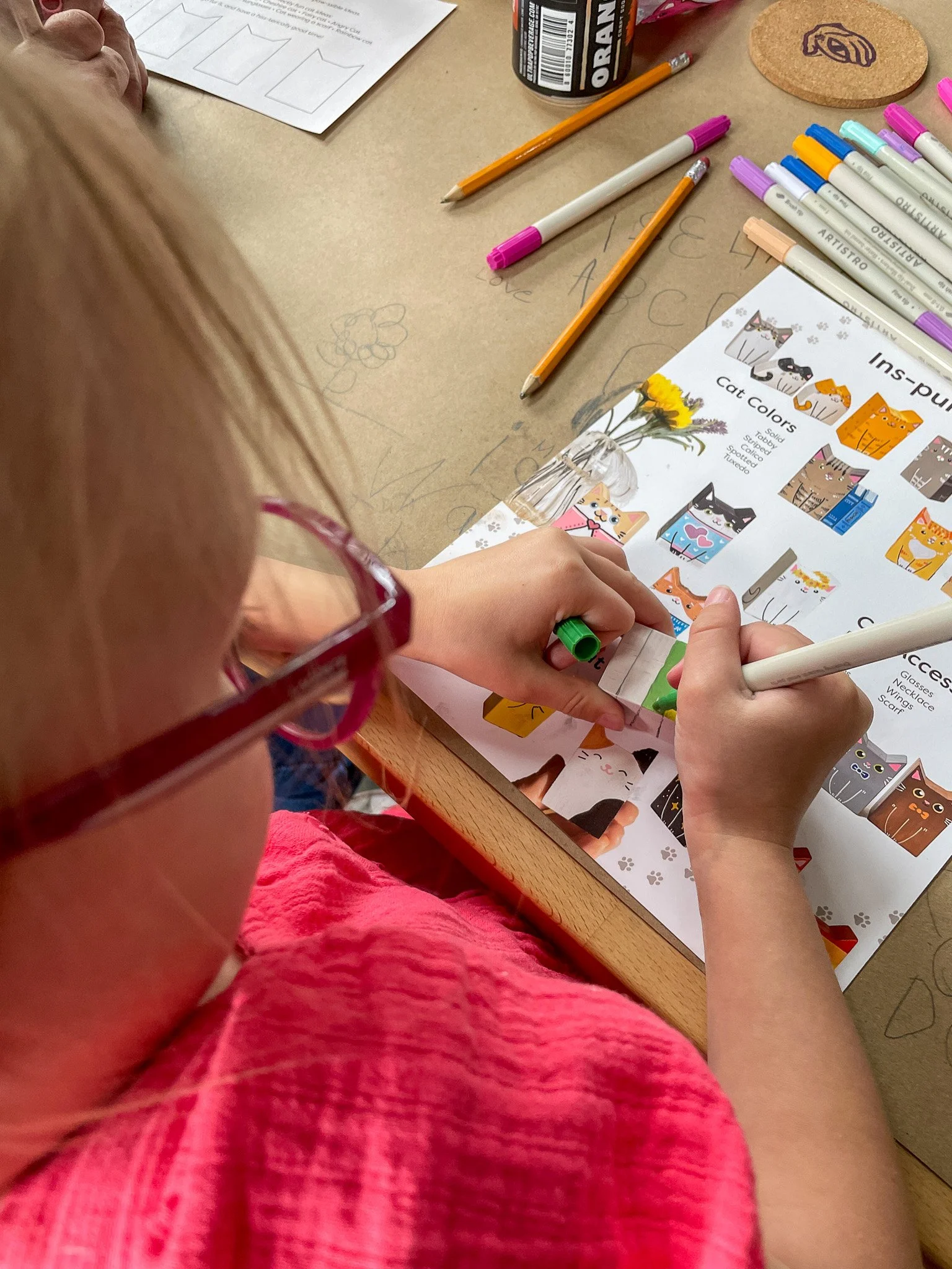 A young girl with red glasses and a pink shirt colors cat stickers from a sticker book labeled 'Cat Colors' at a desk with markers, pencils, and paper.