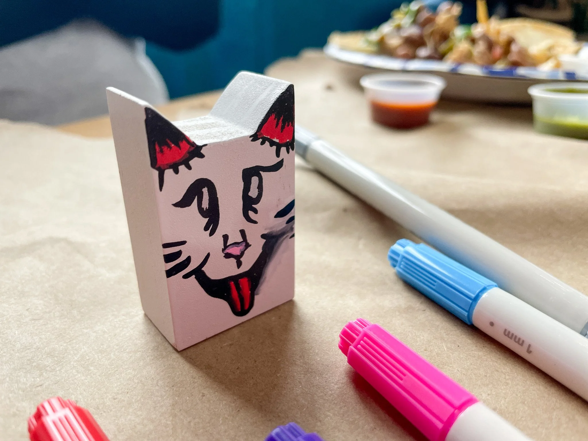 Closeup of a painted cat figurine. Fun wicked face on one side with the tongue sticking out. Paint pens are sitting on the table near it with food in the background