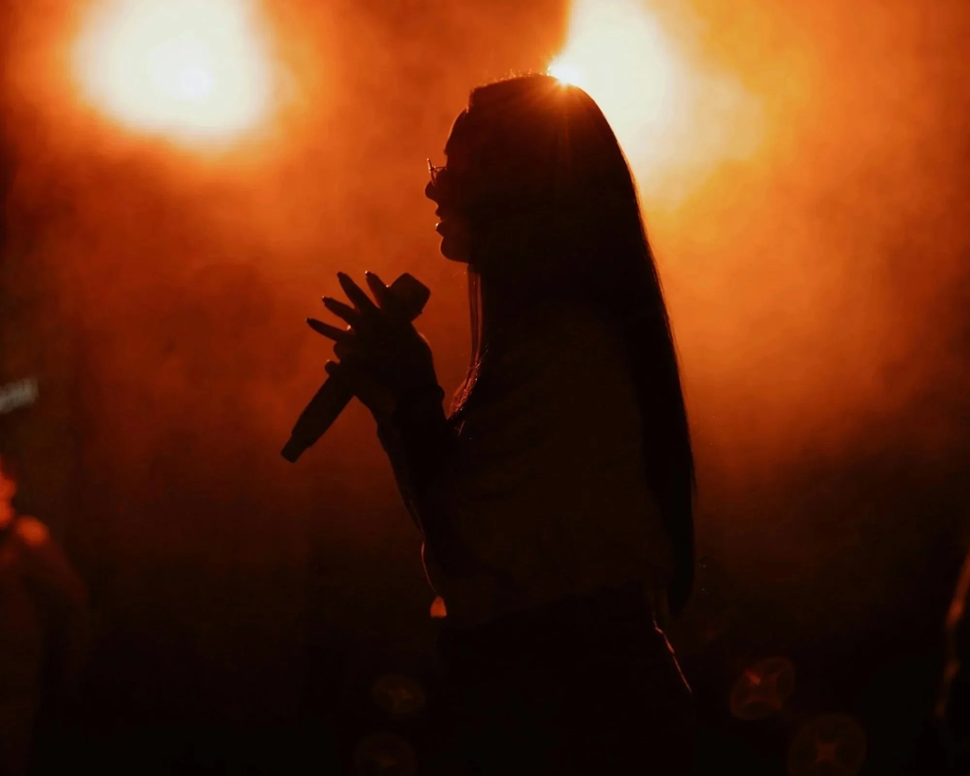 Silhouette of a woman singing into a microphone against a bright, orange-hued background.