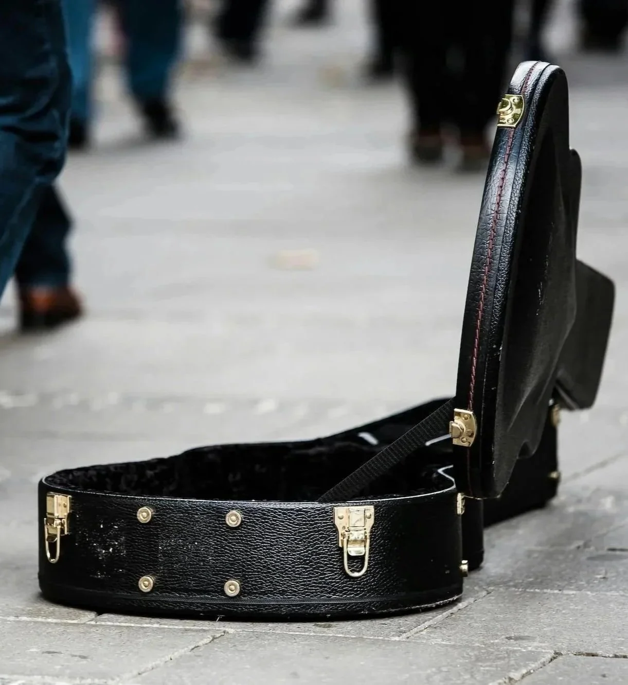 An open black guitar case on a city sidewalk with people walking in the background.