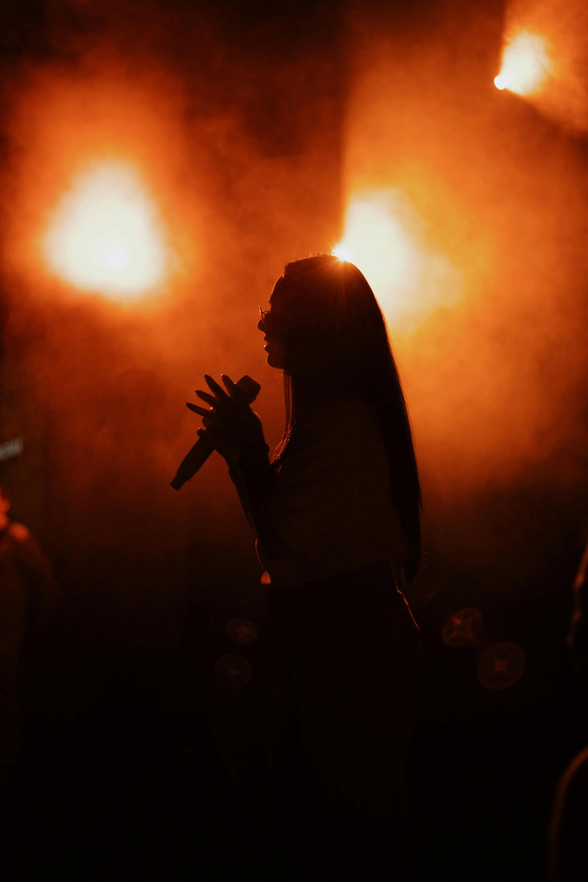 Silhouette of a woman singing into a microphone against a backdrop of bright orange flames or explosions.