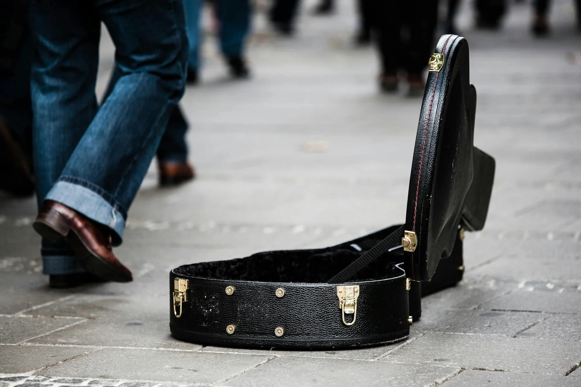 An open black guitar case on a city sidewalk with people walking in the background.