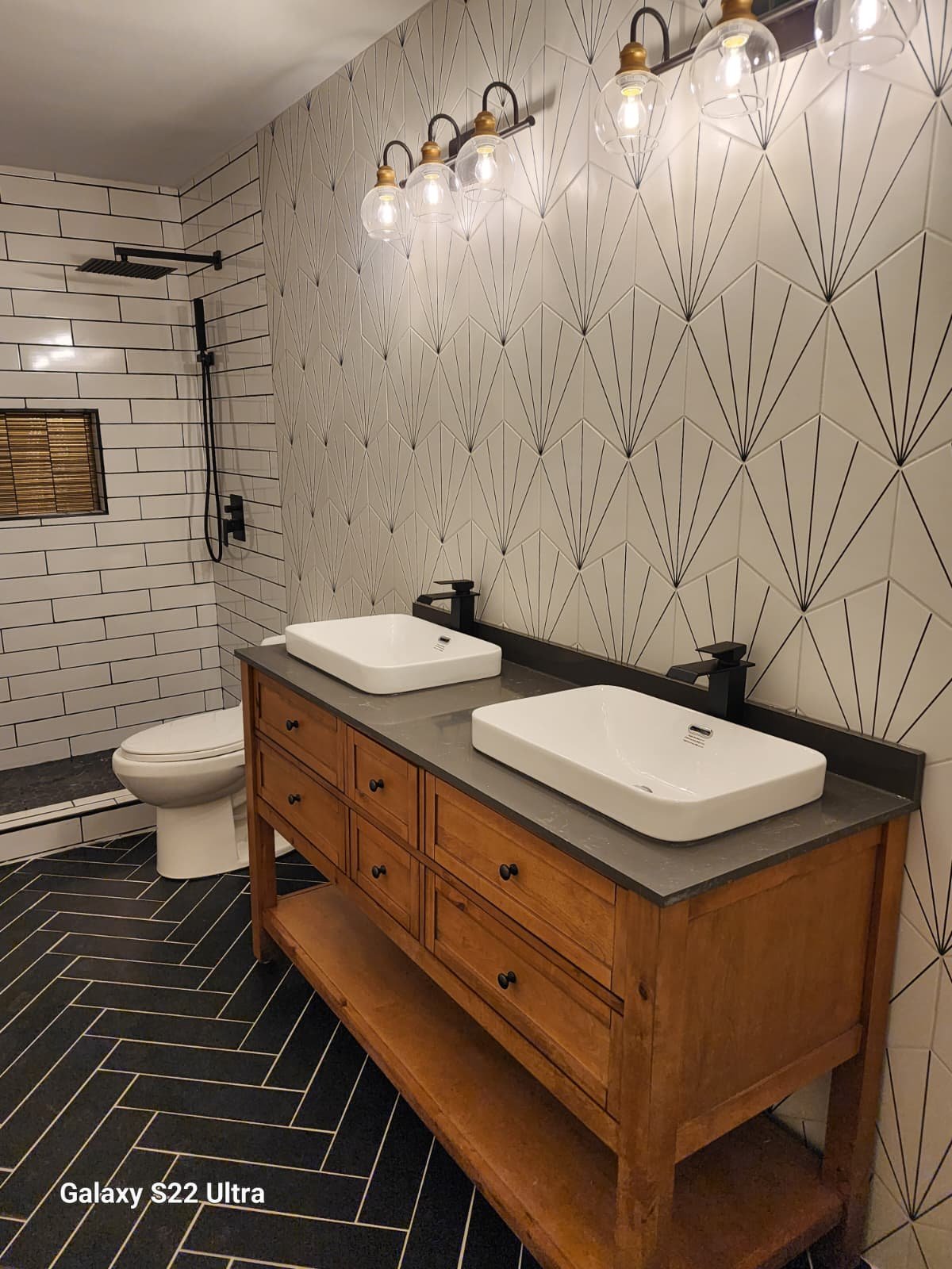 A modern bathroom featuring a double sink vanity with a wooden cabinet, black faucets, and rectangular white sinks. The wall behind the vanity has a geometric patterned tile, and there's a black and gold light fixture above. In the background, a walk-in shower with black fixtures, a rain shower head, and a small window with bamboo blinds.