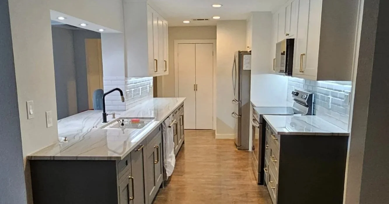 Modern kitchen with white cabinets, marble countertops, stainless steel appliances including a refrigerator and microwave, and a white subway tile backsplash. Hardwood floors and under-cabinet lighting are also visible.