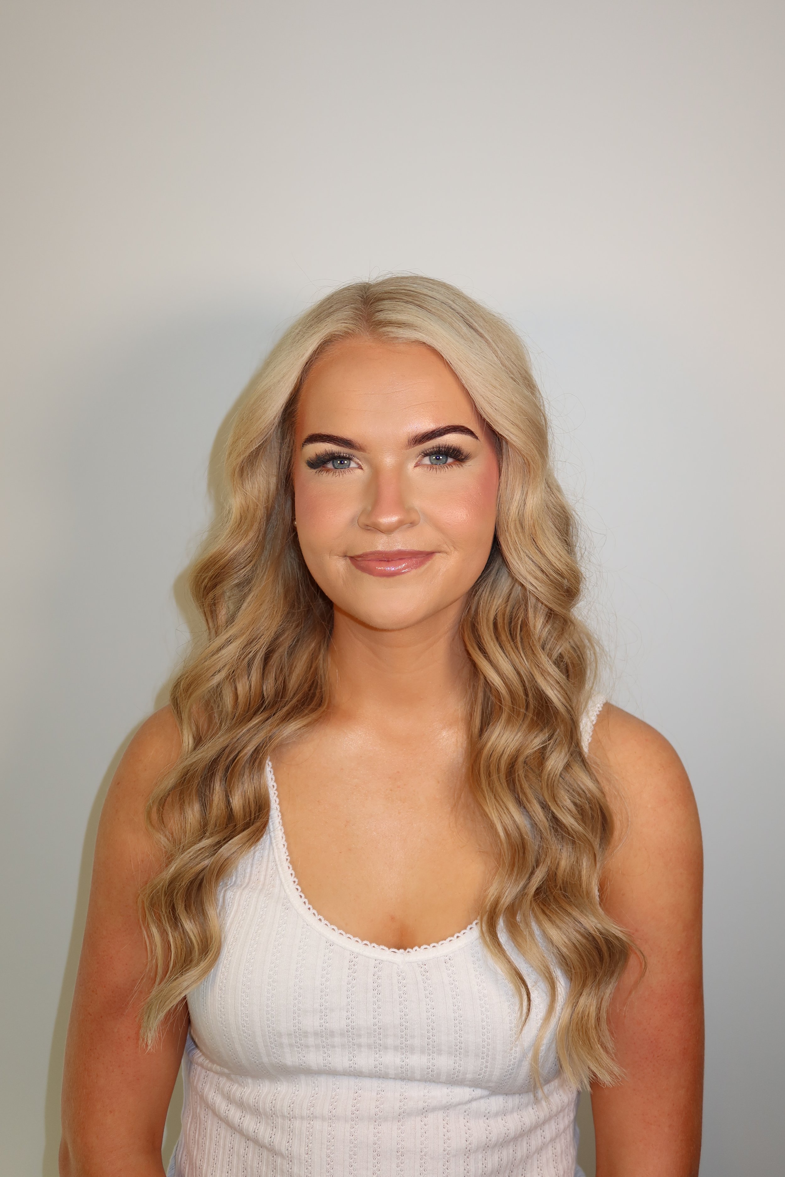 A woman with long, wavy blonde hair and blue eyes, wearing a white sleeveless top, smiling and standing against a plain light-colored wall.