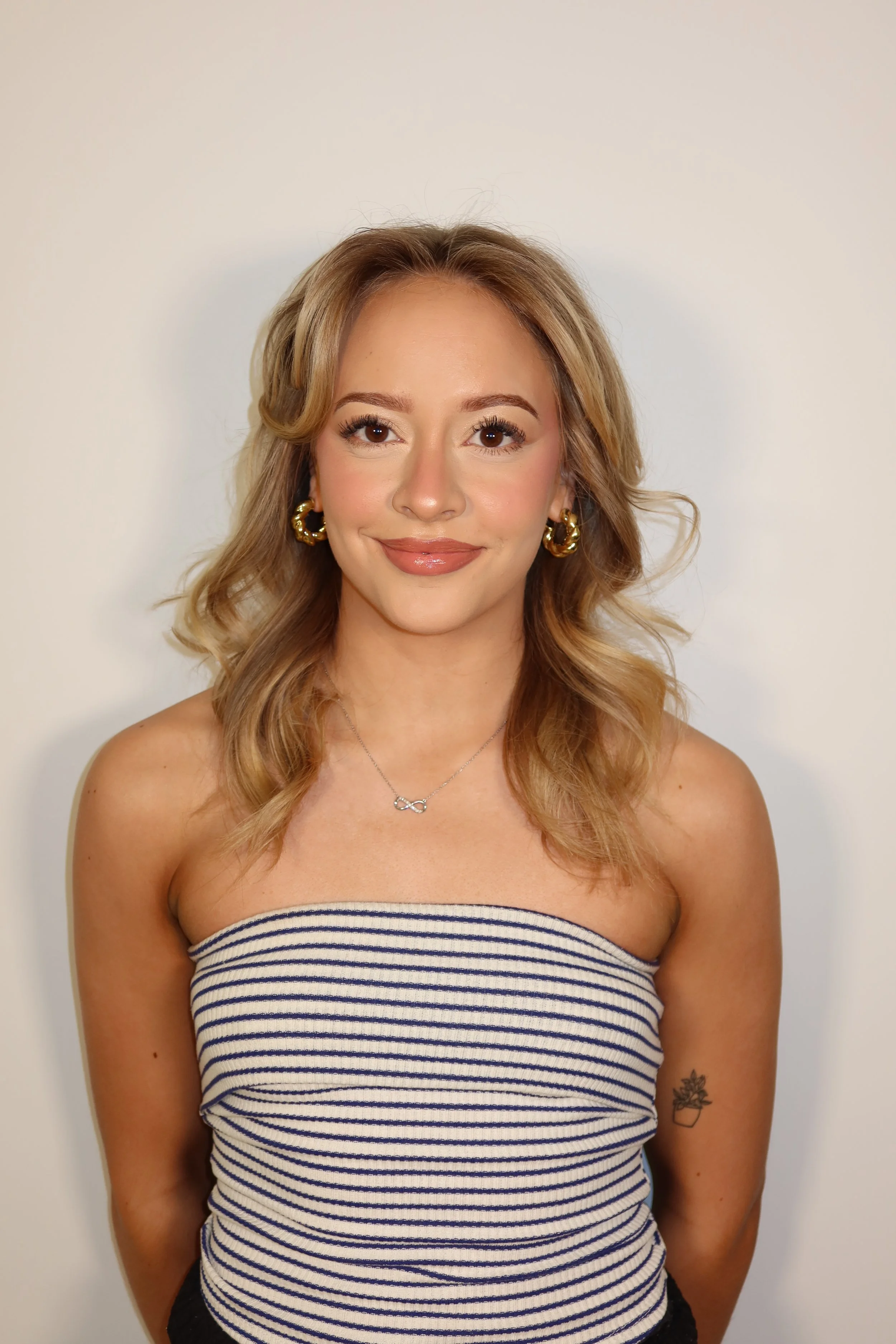 A young woman with wavy blonde hair wearing a striped off-shoulder top, gold hoop earrings, a silver infinity necklace, and a small tattoo of a potted plant on her left arm, standing in front of a plain white wall and smiling at the camera.