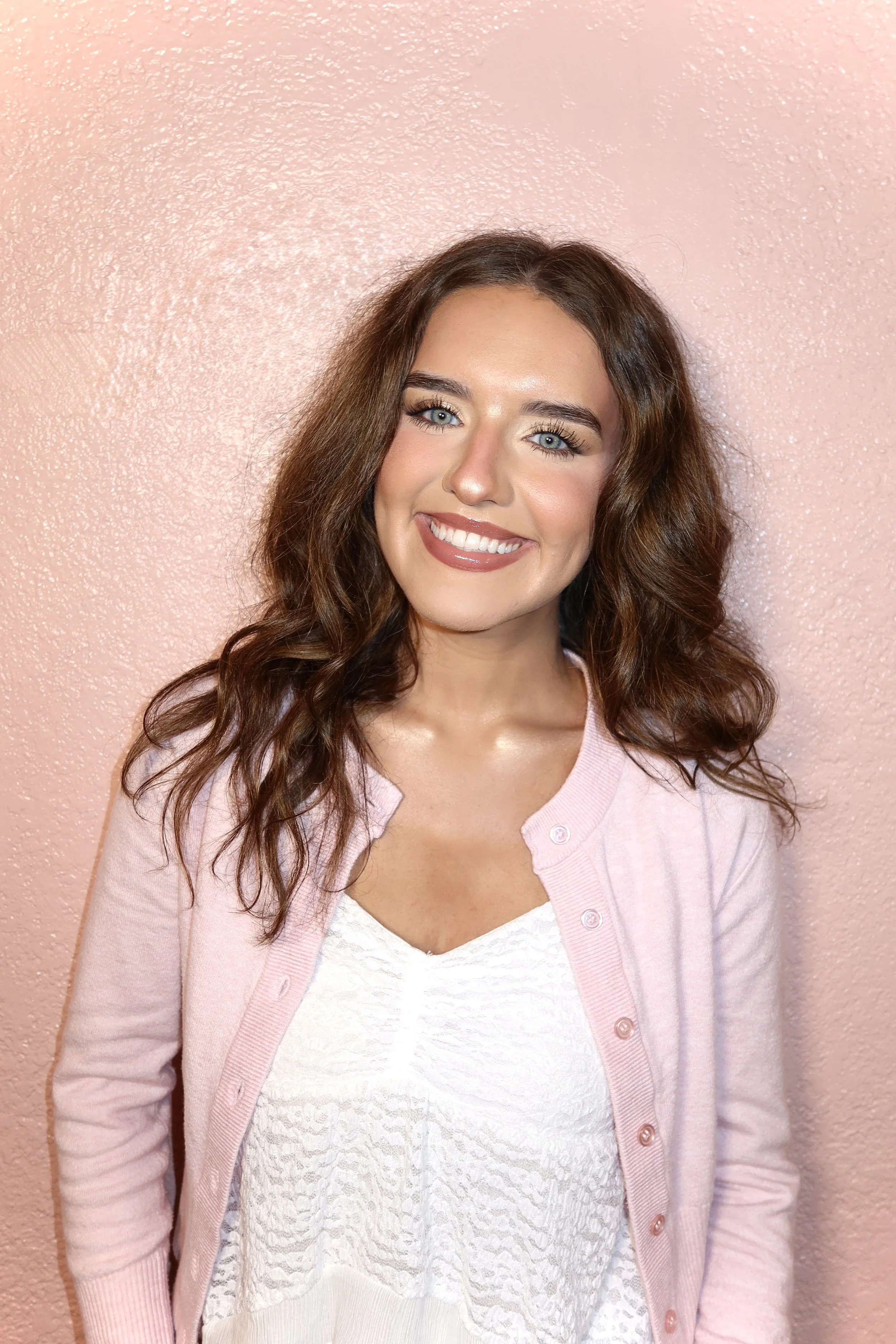 A woman with curly brown hair and blue eyes smiling, wearing a white dress and a pink cardigan, standing against a pink textured wall.