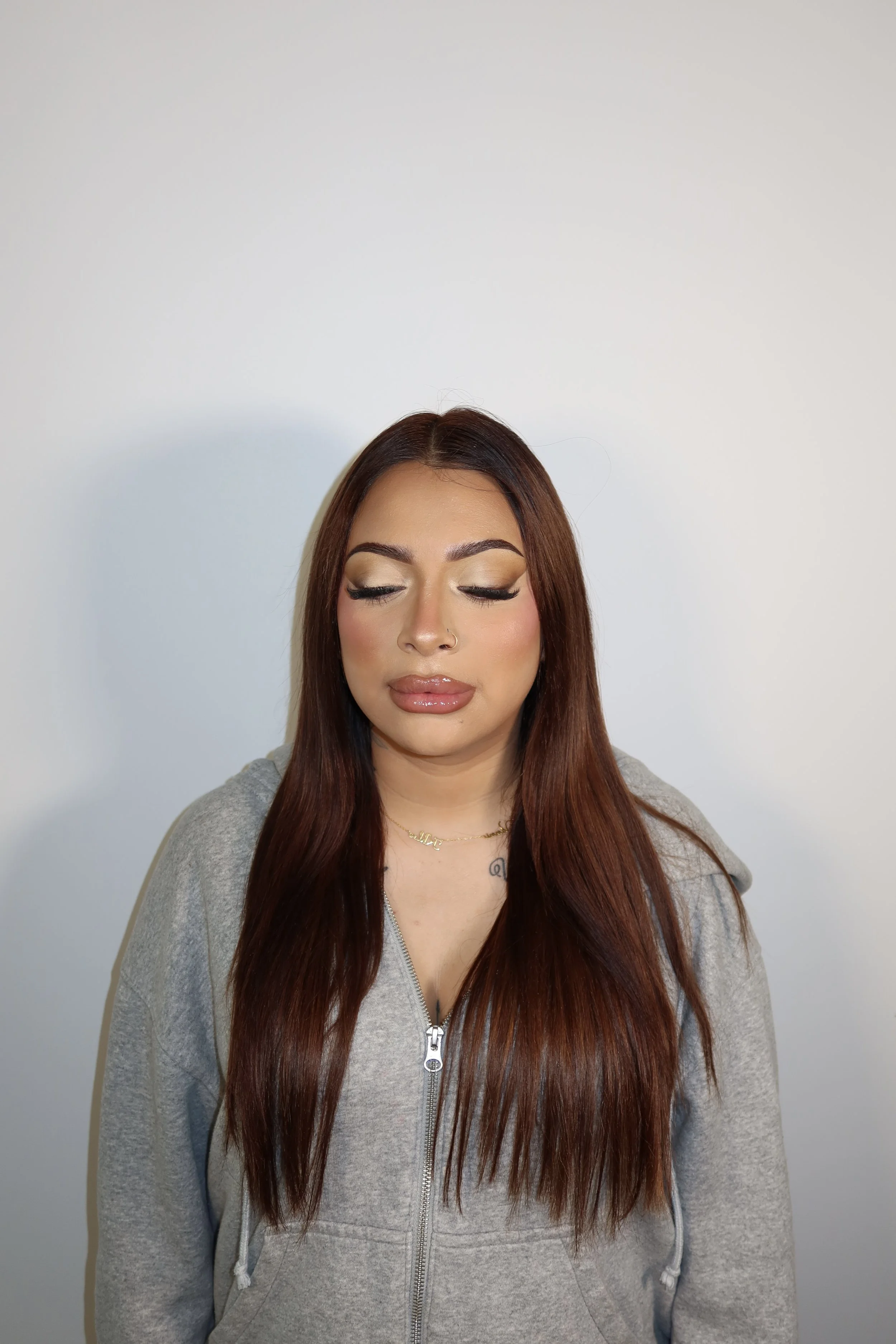 A woman with long brown hair and makeup, wearing a gray zip-up hoodie and a gold necklace, standing against a plain white wall, looking down.