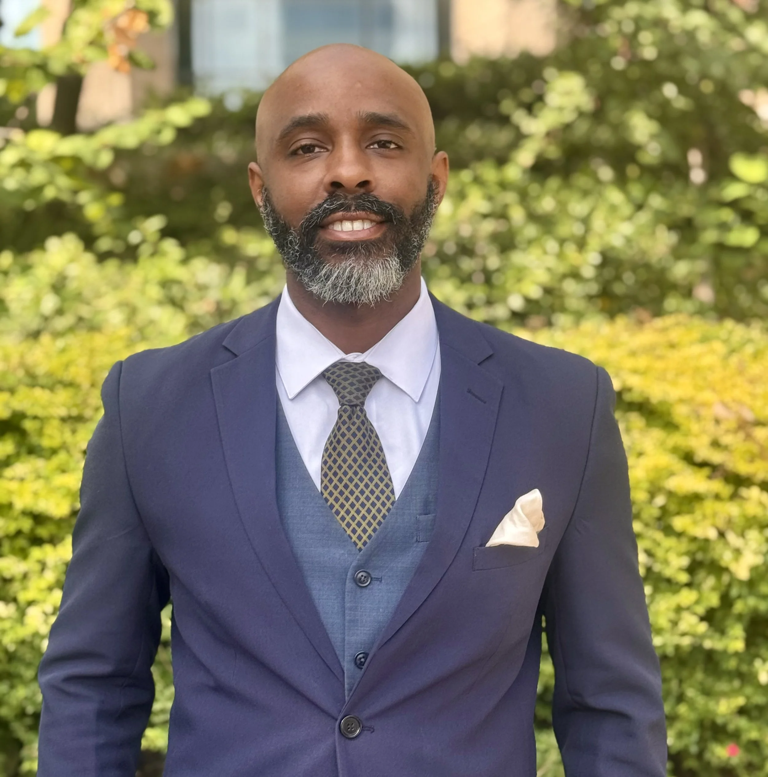 A man with a beard in a navy suit standing outdoors with green plants in the background.