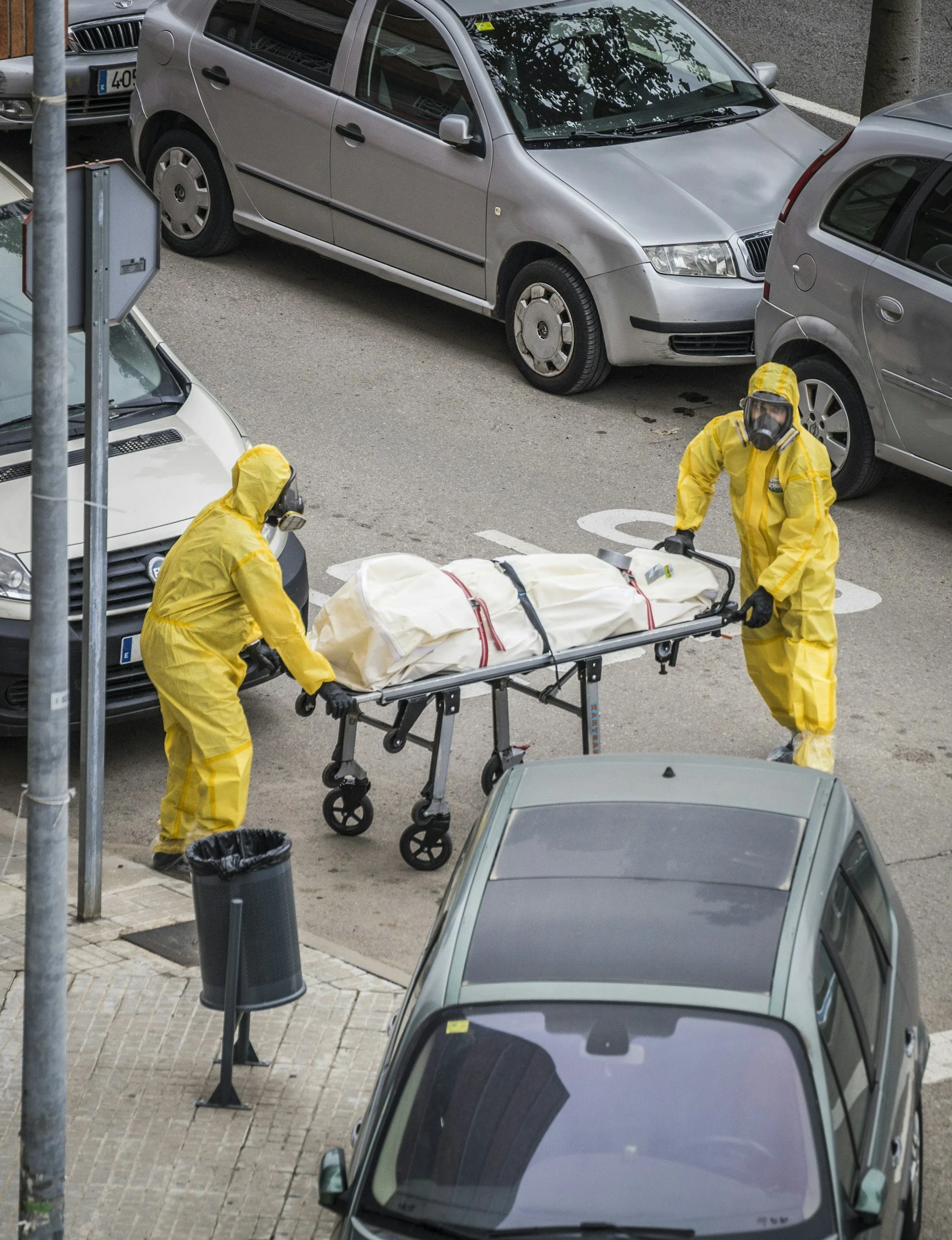 Two individuals dressed in yellow hazmat suits, masks, and gloves are handling a body bag on a stretcher on the street, with multiple parked cars nearby.