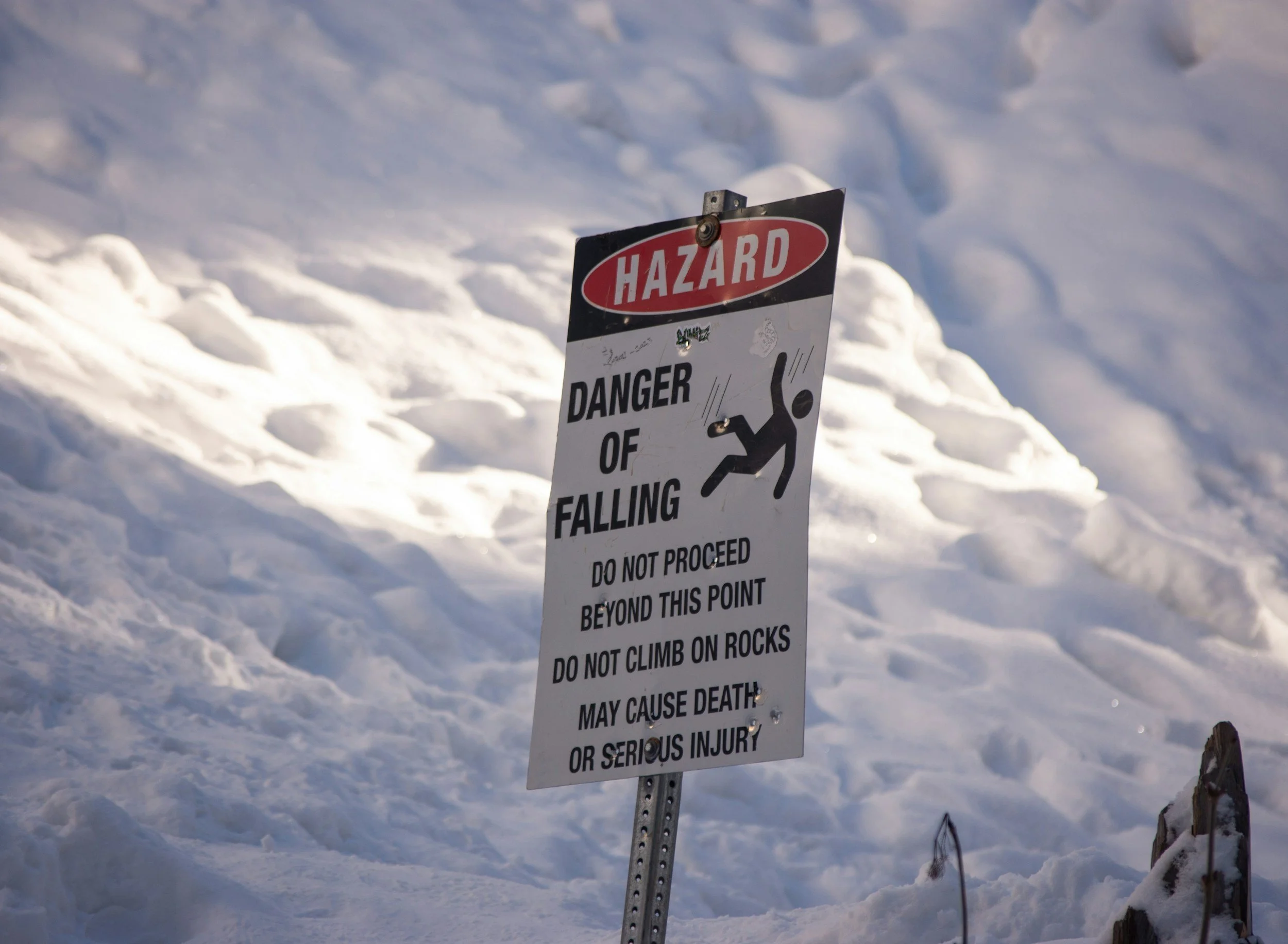 A warning sign on snow-covered terrain cautioning about the danger of falling, prohibiting proceeding beyond this point, climbing on rocks, with a graphic illustration of a person falling.