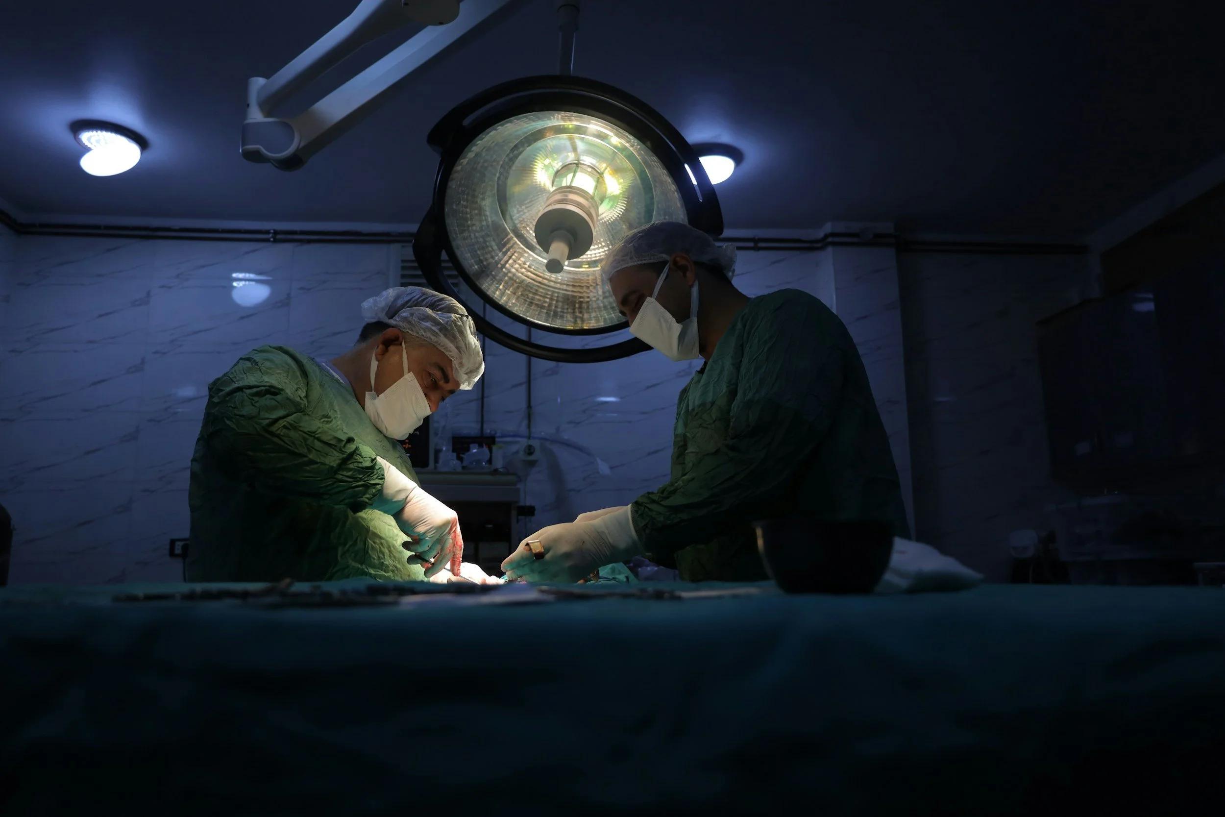 Two medical professionals performing surgery in an operating room, under bright surgical lights, wearing masks and scrubs.