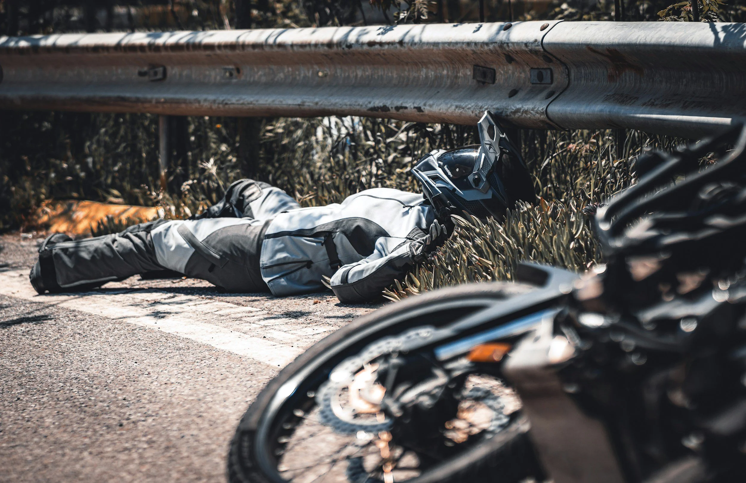 A motorcycle rider in motorcycle gear lying on the ground beside a guardrail, with a helmet on the ground nearby, and a motorcycle partially visible in the foreground.