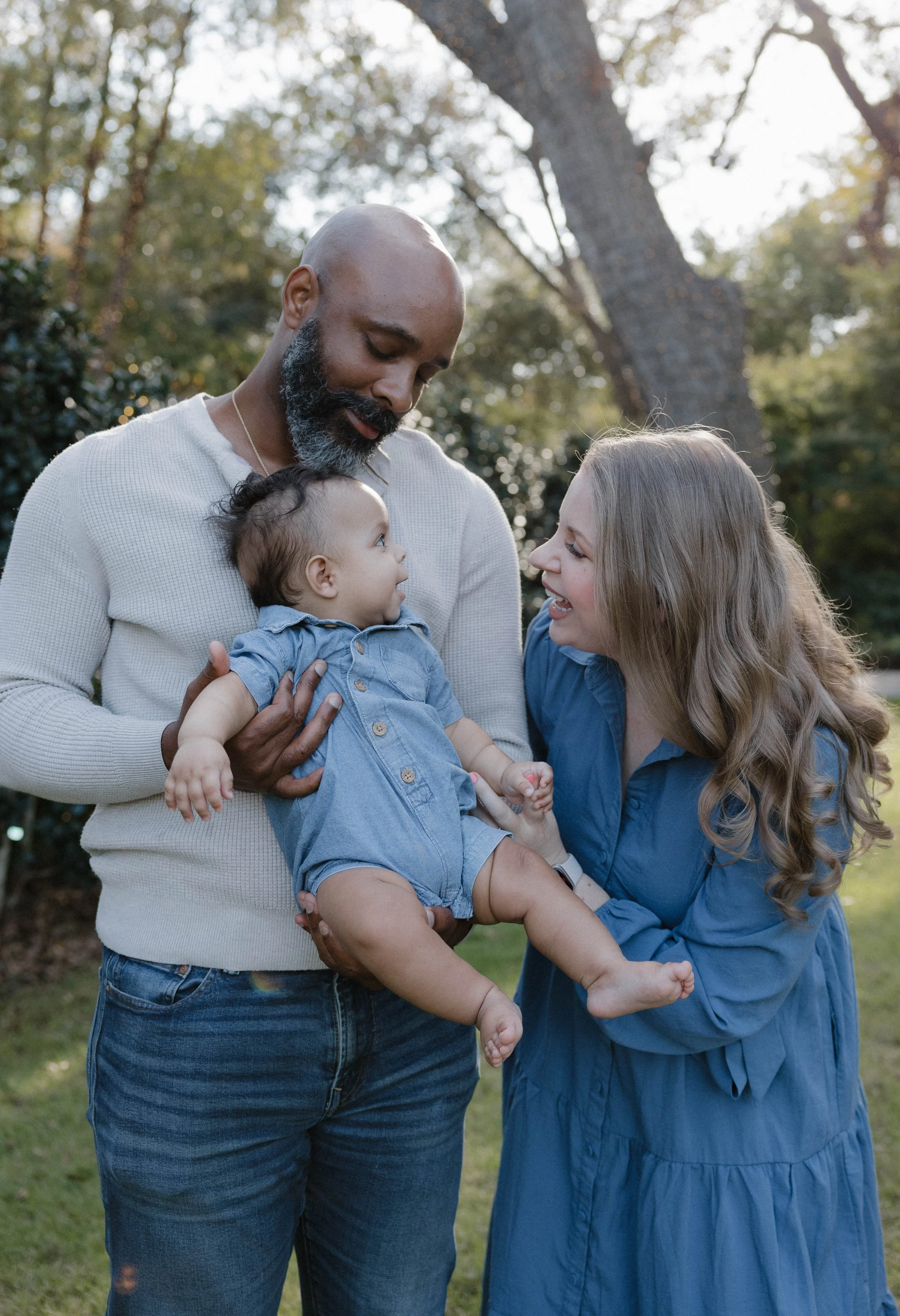 A happy family outdoors in a park during daytime, with a bald man with a beard holding a baby girl in his arms, while a woman with long, wavy hair and a blue dress smiles at the baby. Trees and greenery are visible in the background.