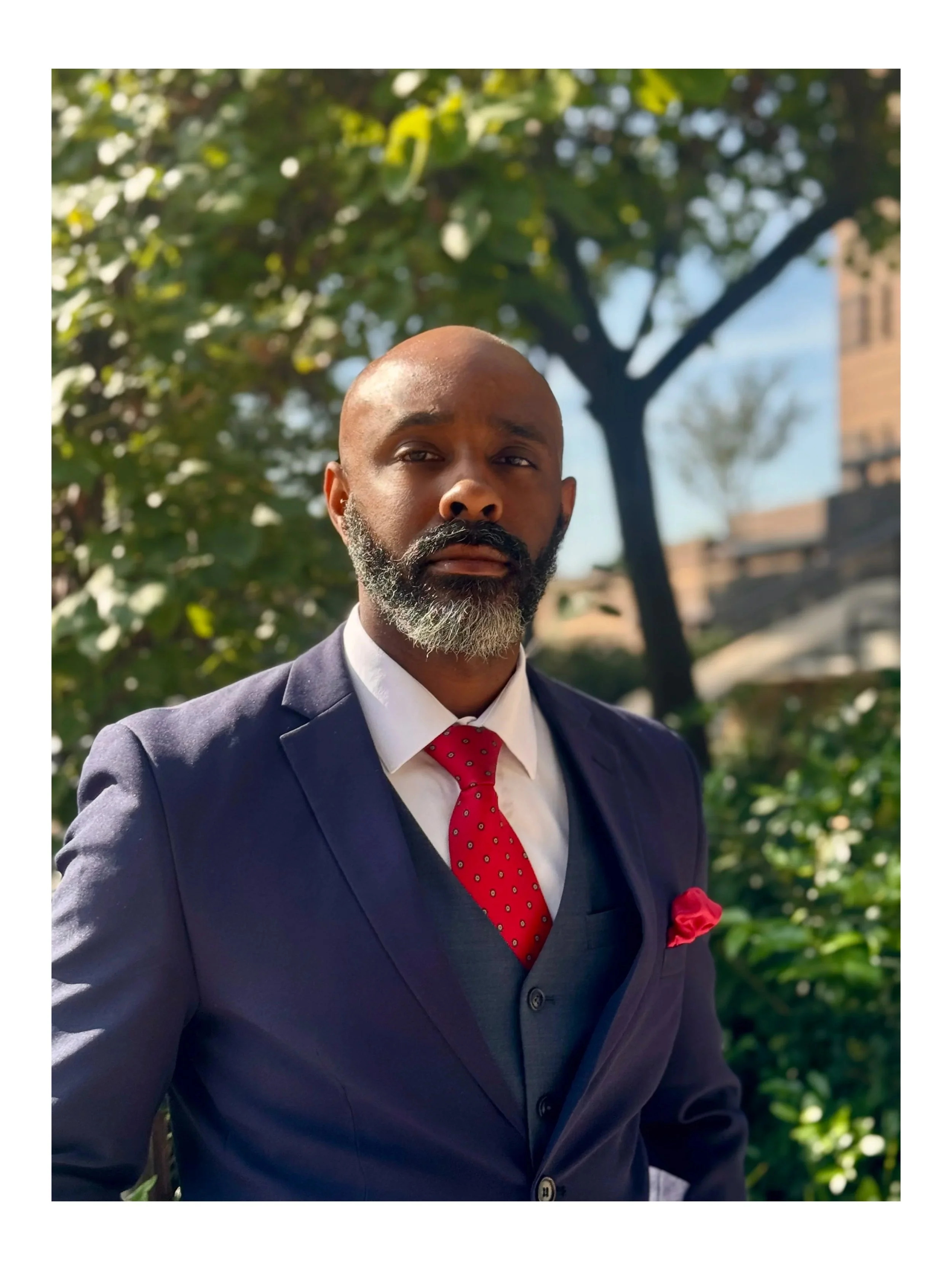 A man in a dark suit with a red tie and pocket square standing outdoors with trees and a building in the background.