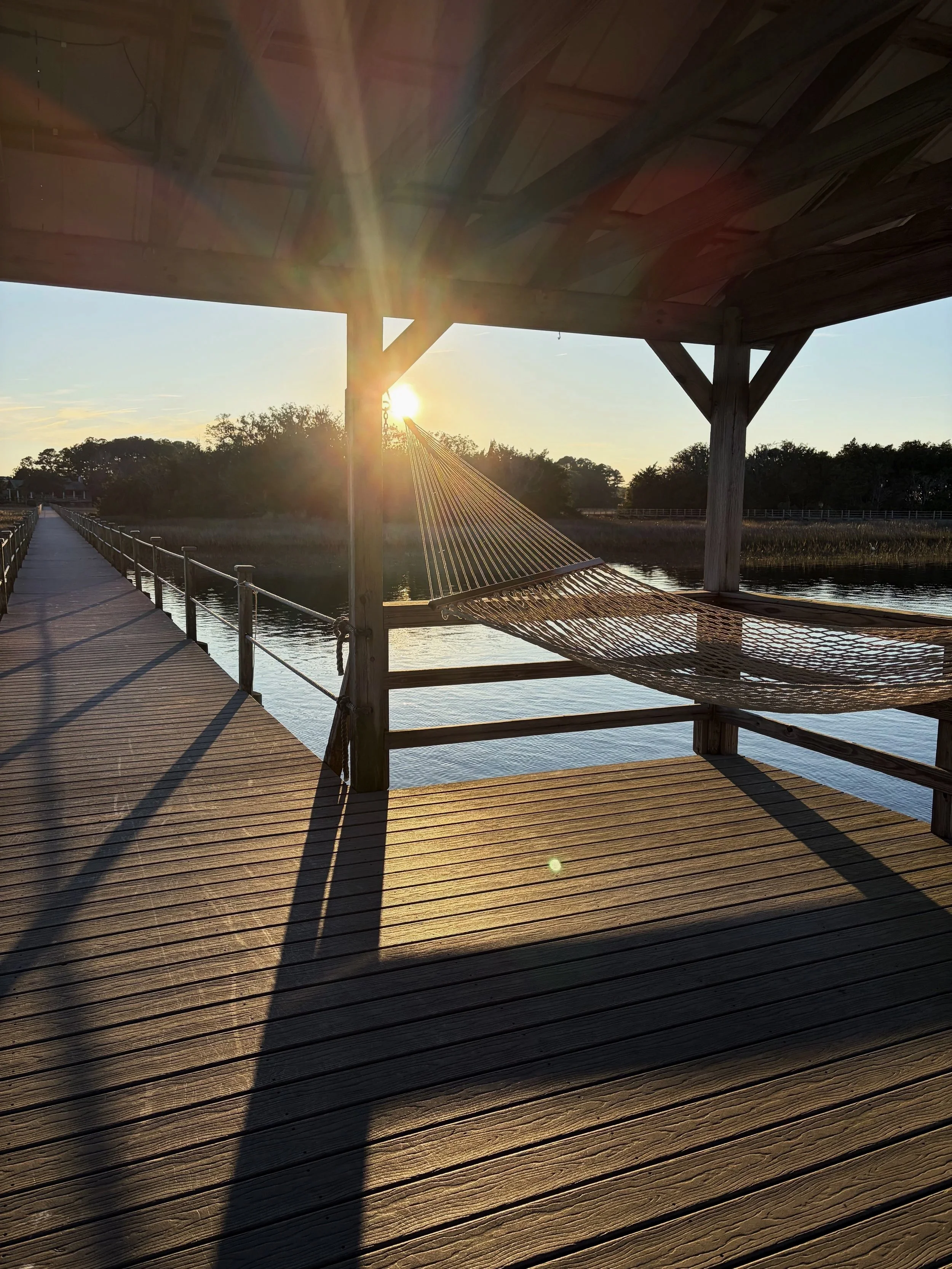 Sunset view on a wooden dock overlooking water, with a hammock under a covered area and trees in the distance.