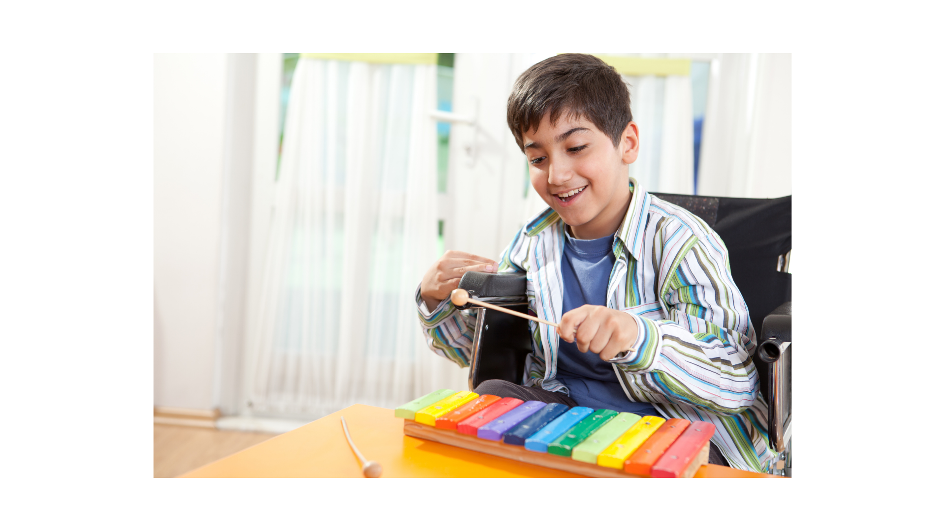 A boy in a wheelchair playing a colorful xylophone with a mallet, smiling indoors near a window with white curtains.