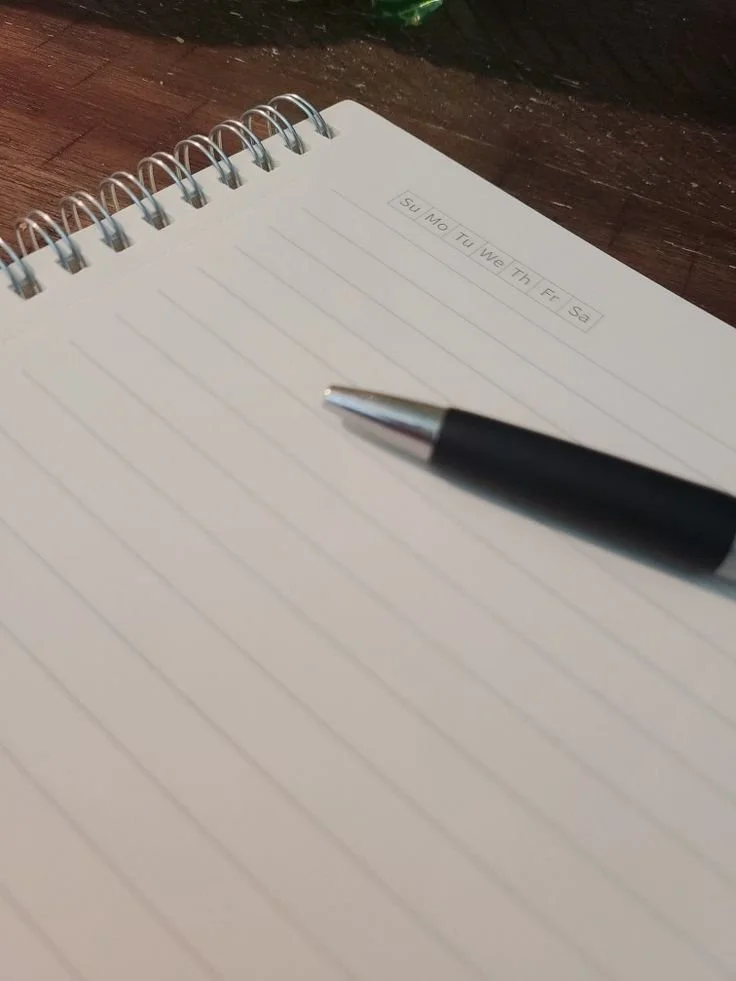Open lined notebook with a black pen resting on it, showing days of the week at the top