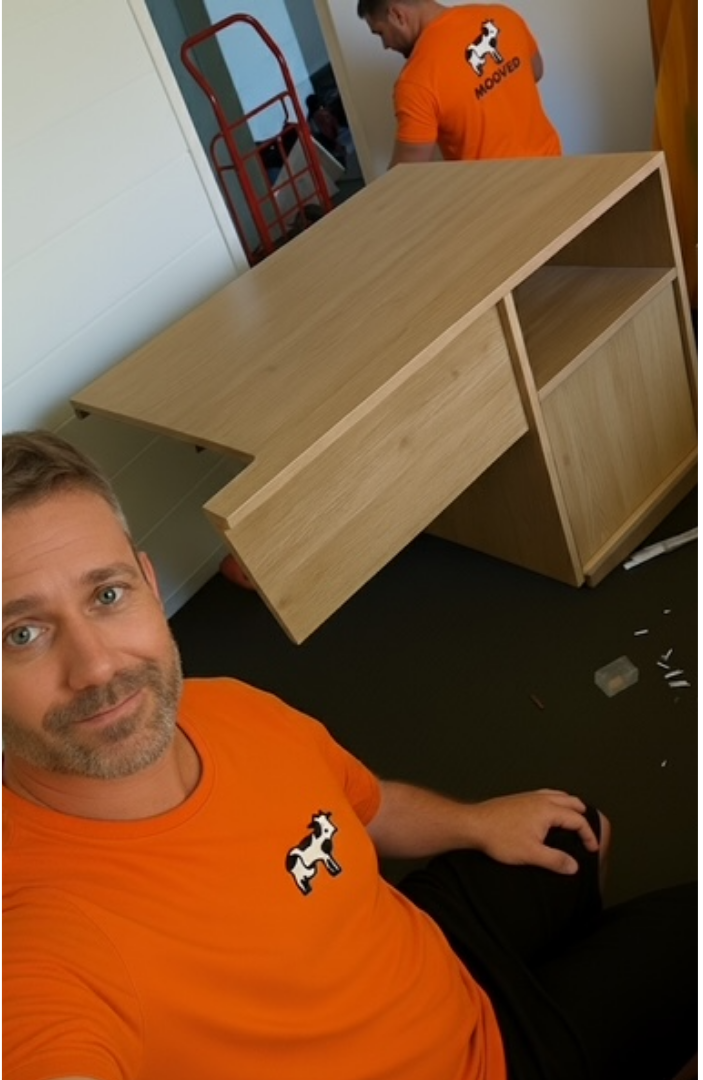 A man taking a selfie sitting on the floor, wearing an orange shirt with a cartoon dog mascot on it, in front of a partially assembled wooden desk.