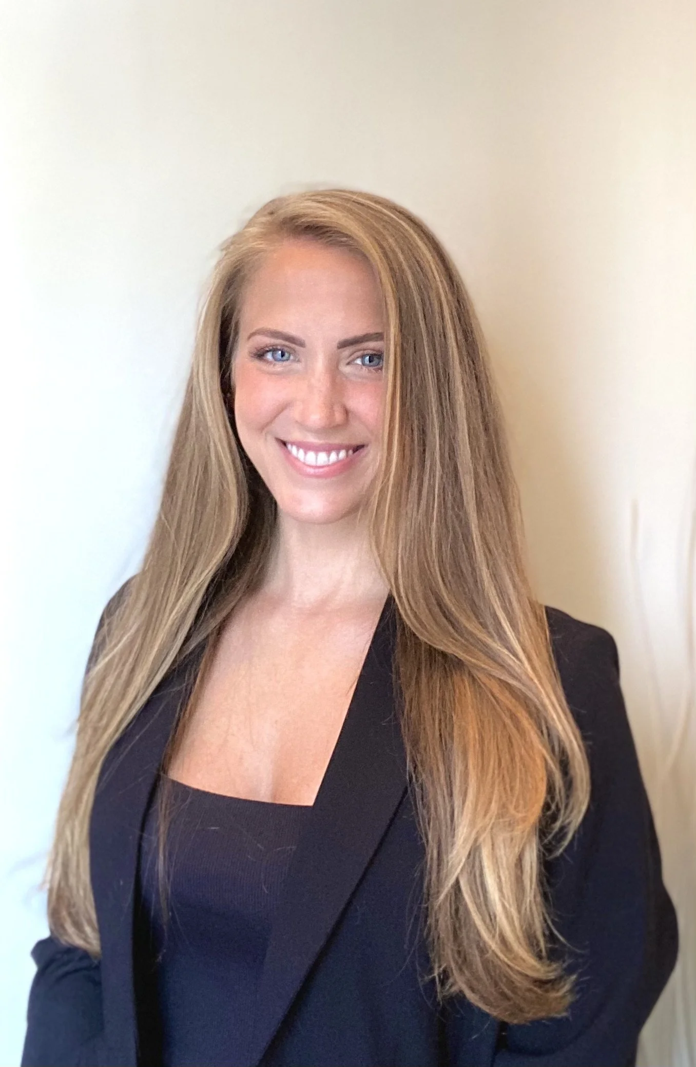 A woman with long, straight blonde hair, blue eyes, and a bright smile, wearing a black blazer, standing against a plain beige wall.