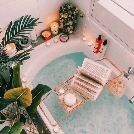 Bathtub filled with water, surrounded by candles, plants, and toiletries, with an open book and bath accessories on a tray resting on the edge.