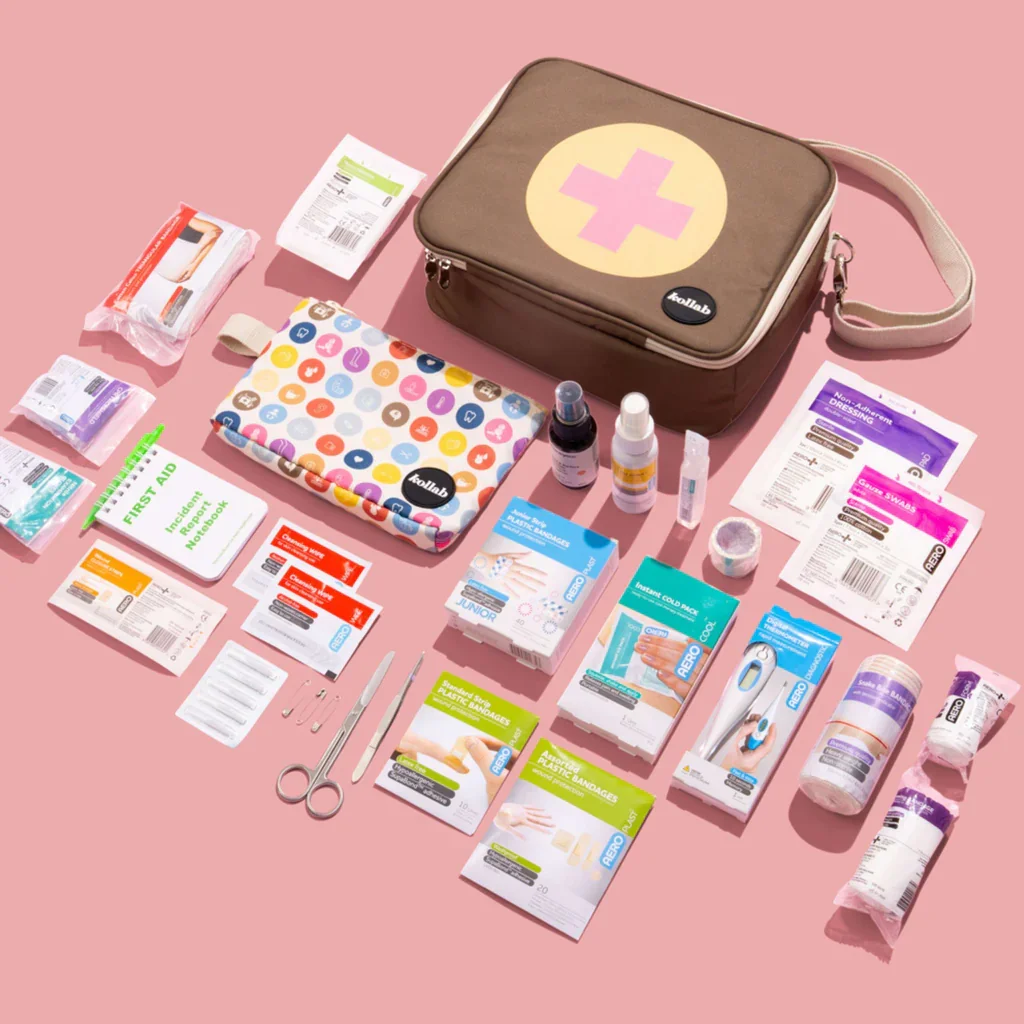 First aid kit with various medical supplies such as bandages, antiseptic wipes, scissors, thermometer, and medications laid out on a pink background.