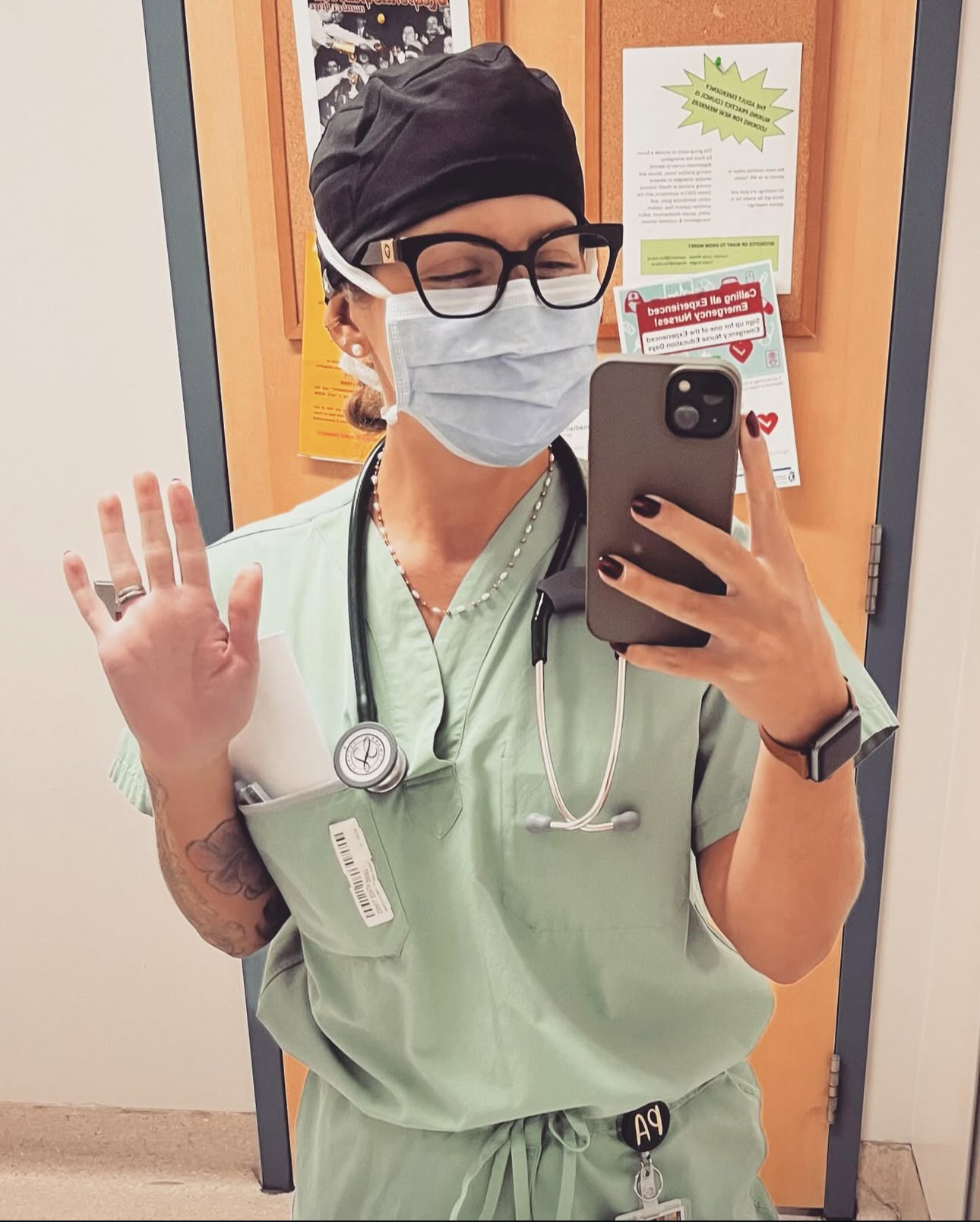 A nurse taking a selfie in a mirror, wearing green scrubs, a black cap, glasses, a face mask, and a stethoscope around her neck. She is in a hospital or clinic hallway, with notices on the door behind her.