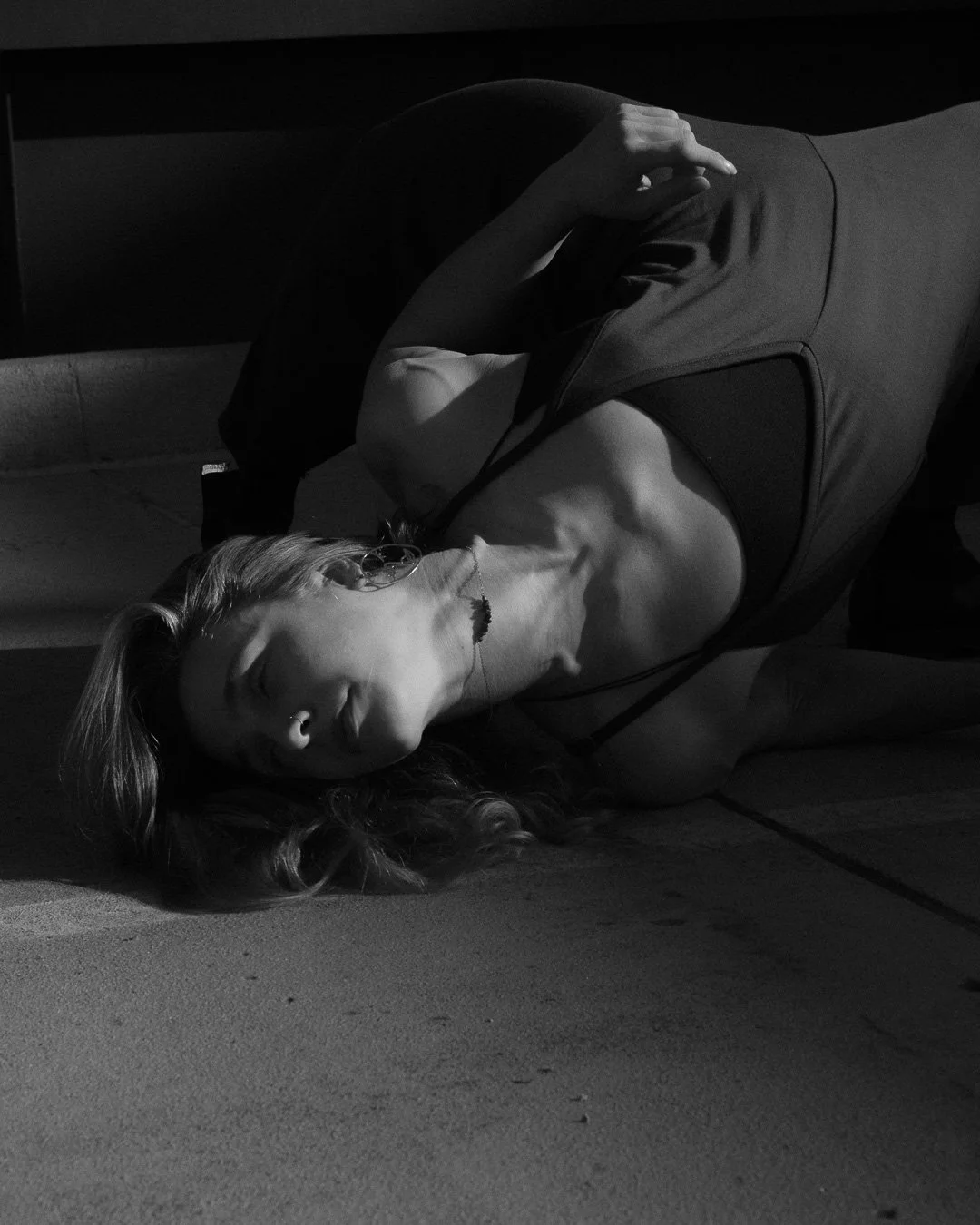 A woman with long hair lying on the floor with her eyes closed, resting her head on the ground. She is wearing a sleeveless top and a necklace, with one hand resting on her forehead. The photo is in black and white with dramatic lighting.