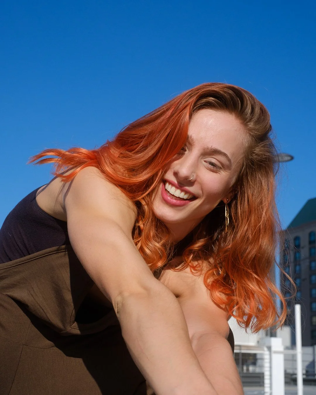 A young woman with red hair and a nose piercing, smiling with her eyes closed, leaning forward outdoors against a backdrop of blue sky and a cityscape.
