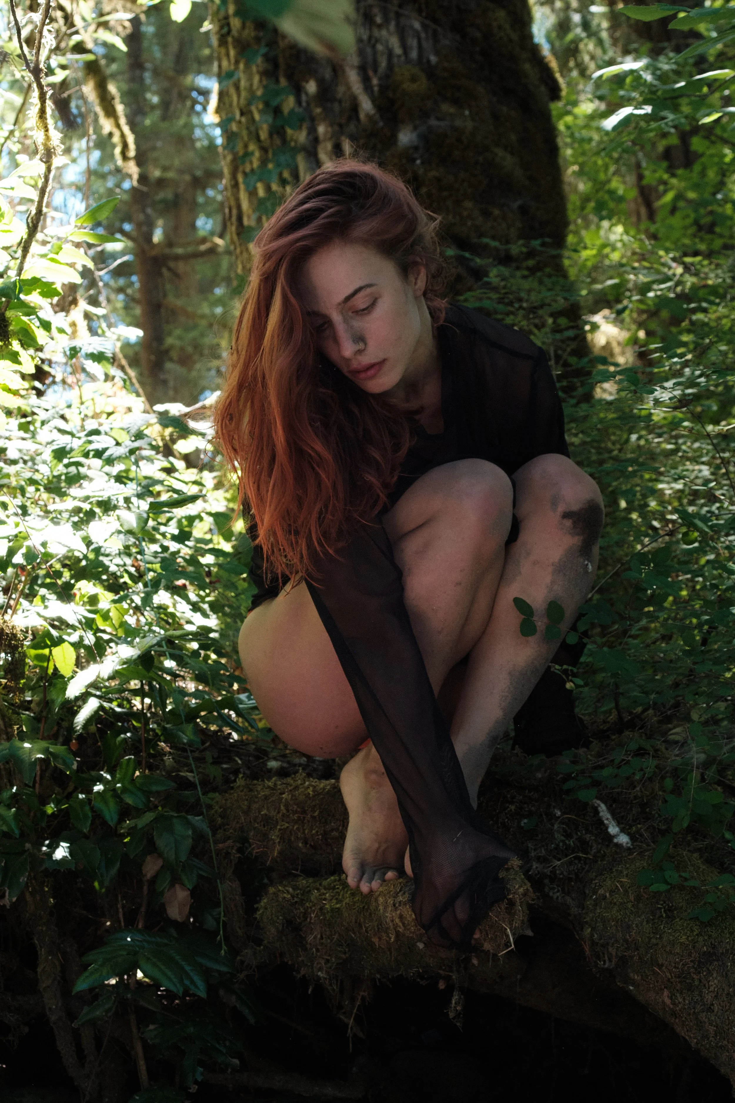A woman with long red hair crouches on a mossy log in a forest, surrounded by green foliage and sunlight filtering through the trees.
