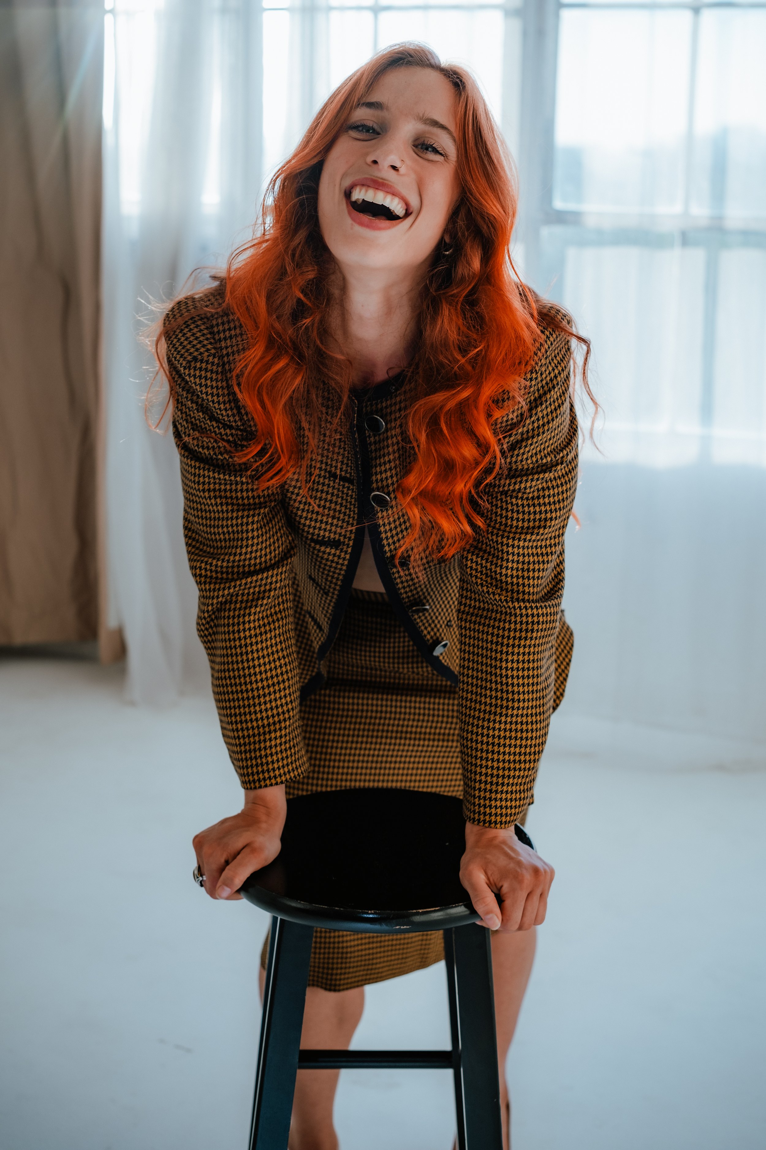 A woman with red hair laughing and leaning forward on a black stool in a bright room with large windows.