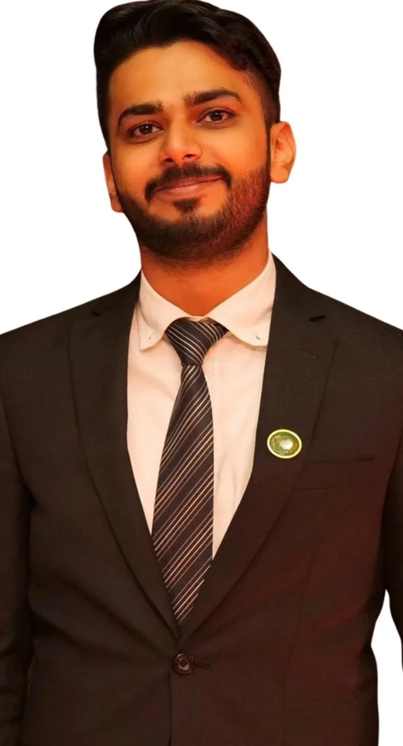 A young man dressed in a black suit, white shirt, and striped tie, with a badge on his lapel, standing against a white background, smiling.