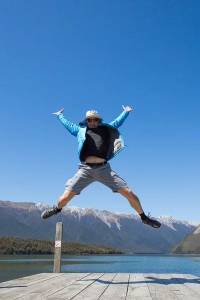 Person jumping on a dock over a lake with mountains in the background under a clear blue sky.