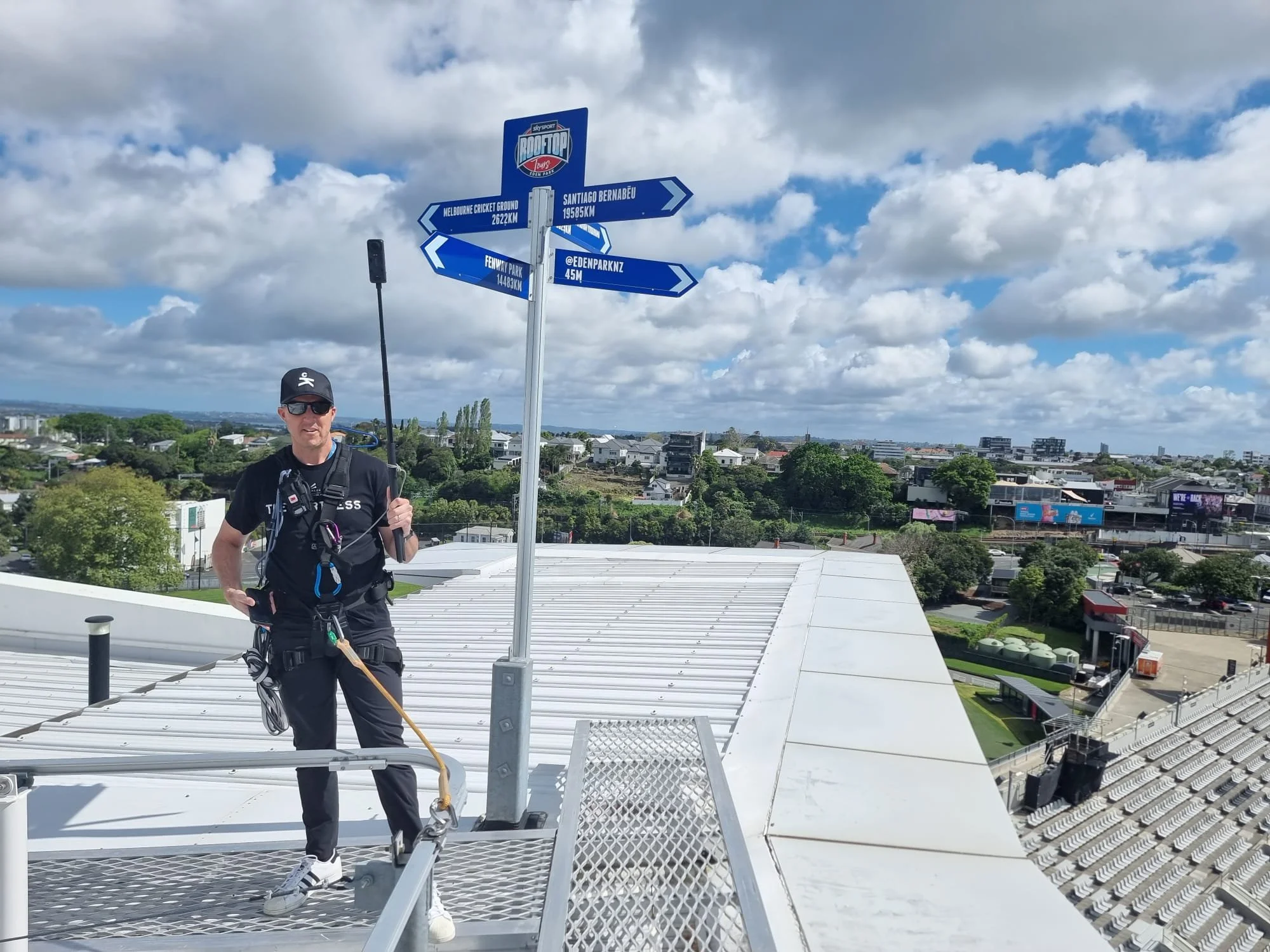 A person standing on an  rooftop of one of the stadiums at Eden Park with safety harnesses, near a signpost with multiple arrows indicating directions and distances to various locations, in a cityscape background under a partly cloudy sky.