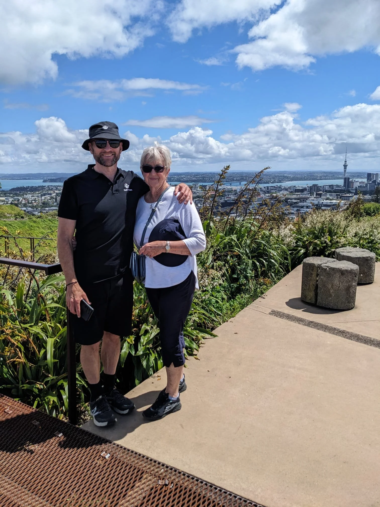 Tour guide and customer on top of Maungawhau/Mt Eden with the Auckland CBD and Waitemata Harbour in the background