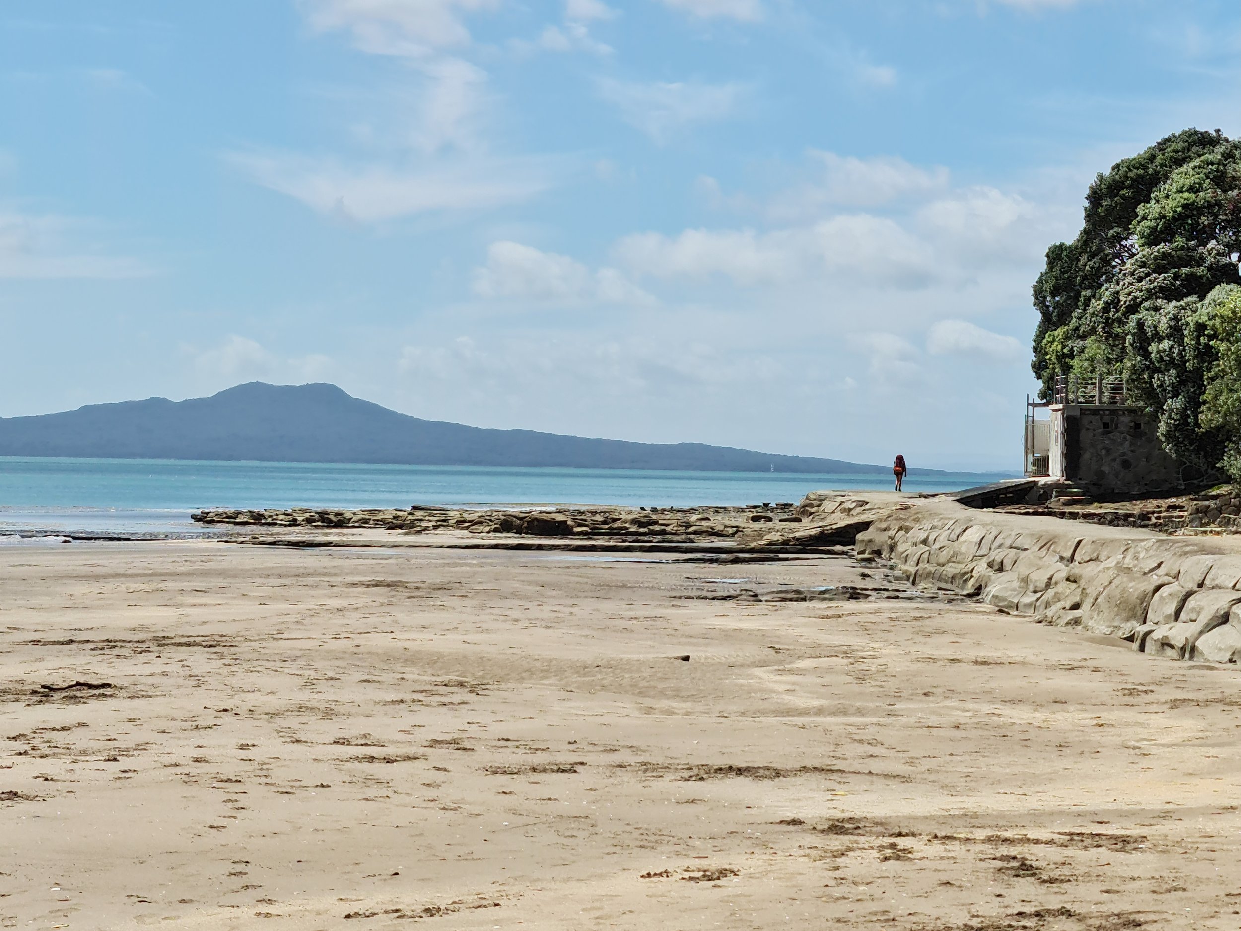 A wide sandy beach with a calm ocean and distant volcanic island in the background. A person with a backpack is hiking the Te Araroa path near a rocky shoreline on the right side, with trees and a small structure nearby. The sky is partly cloudy.